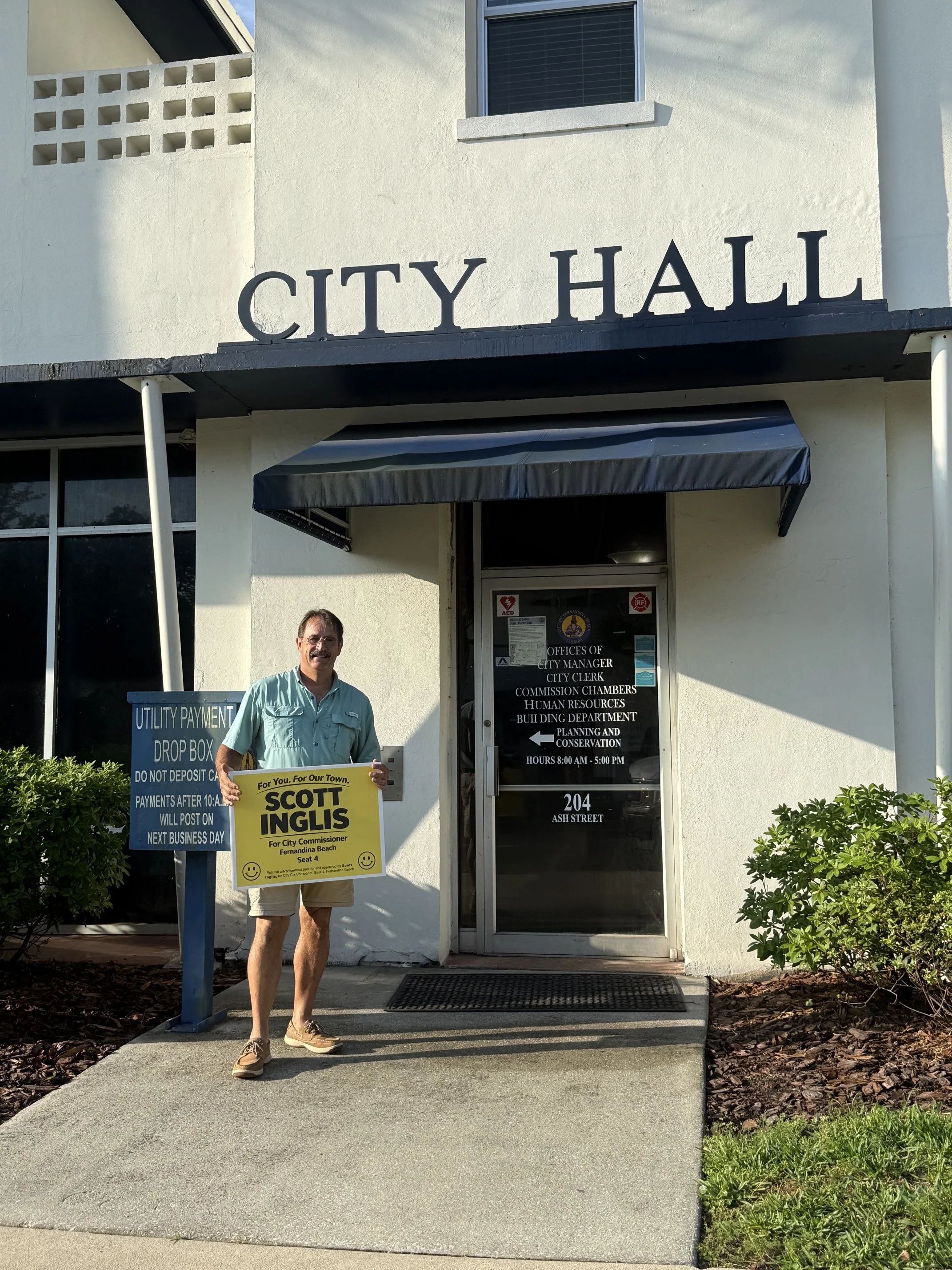 A man standing outside the city hall holding a yellow campaign sign that reads 'For you, for our town. Scott Inglis for City Commissioner Fernandina Beach, Seat 4.' There is a blue sign to the left listing utility payment drop box rules. The building has a white exterior with black lettering spelling 'CITY HALL' above the entrance, which has a black awning and glass door with various notices on it. There are plants and bushes on either side of the entrance.