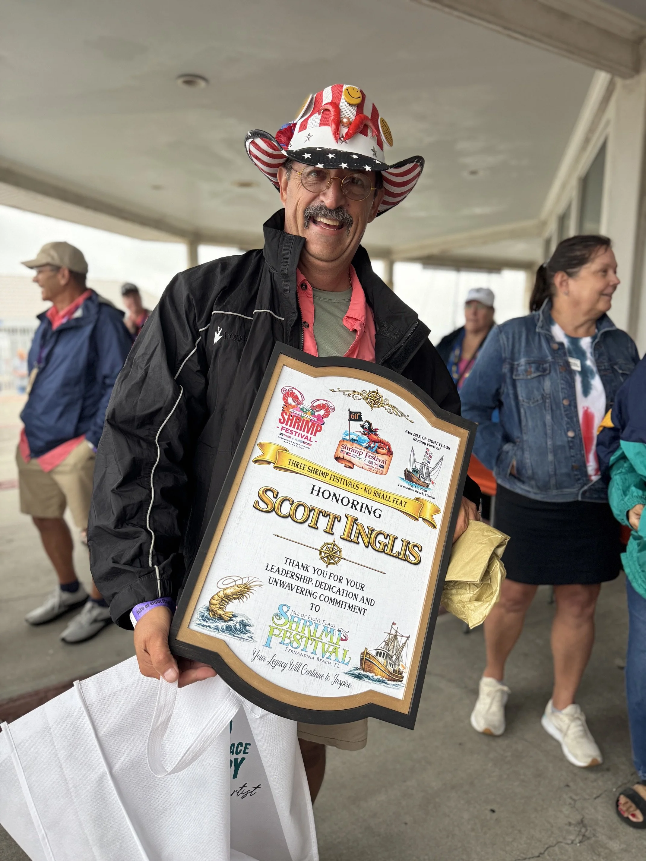 A man wearing a patriotic cowboy hat decorated with smiley faces, stars, and stripes, smiling while holding a framed certificate or award. Several people are visible in the background, standing on an outdoor patio or balcony.