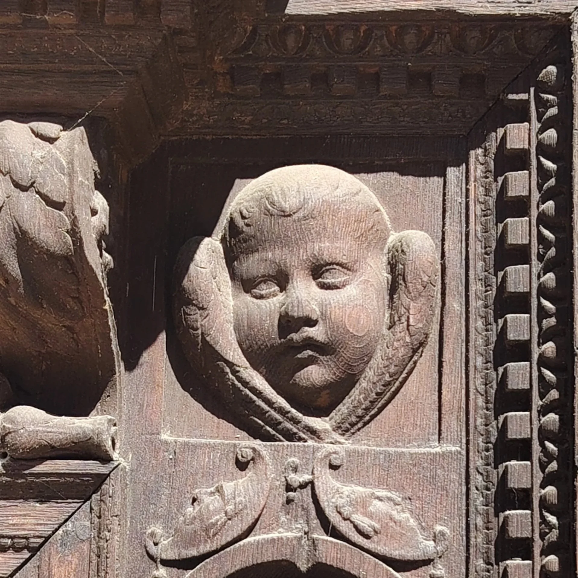Détails d'une sculpture en pierre représentant une tête d'enfant, entourée de corbeaux sculptés, avec des éléments architecturaux en bois ornés en arrière-plan.