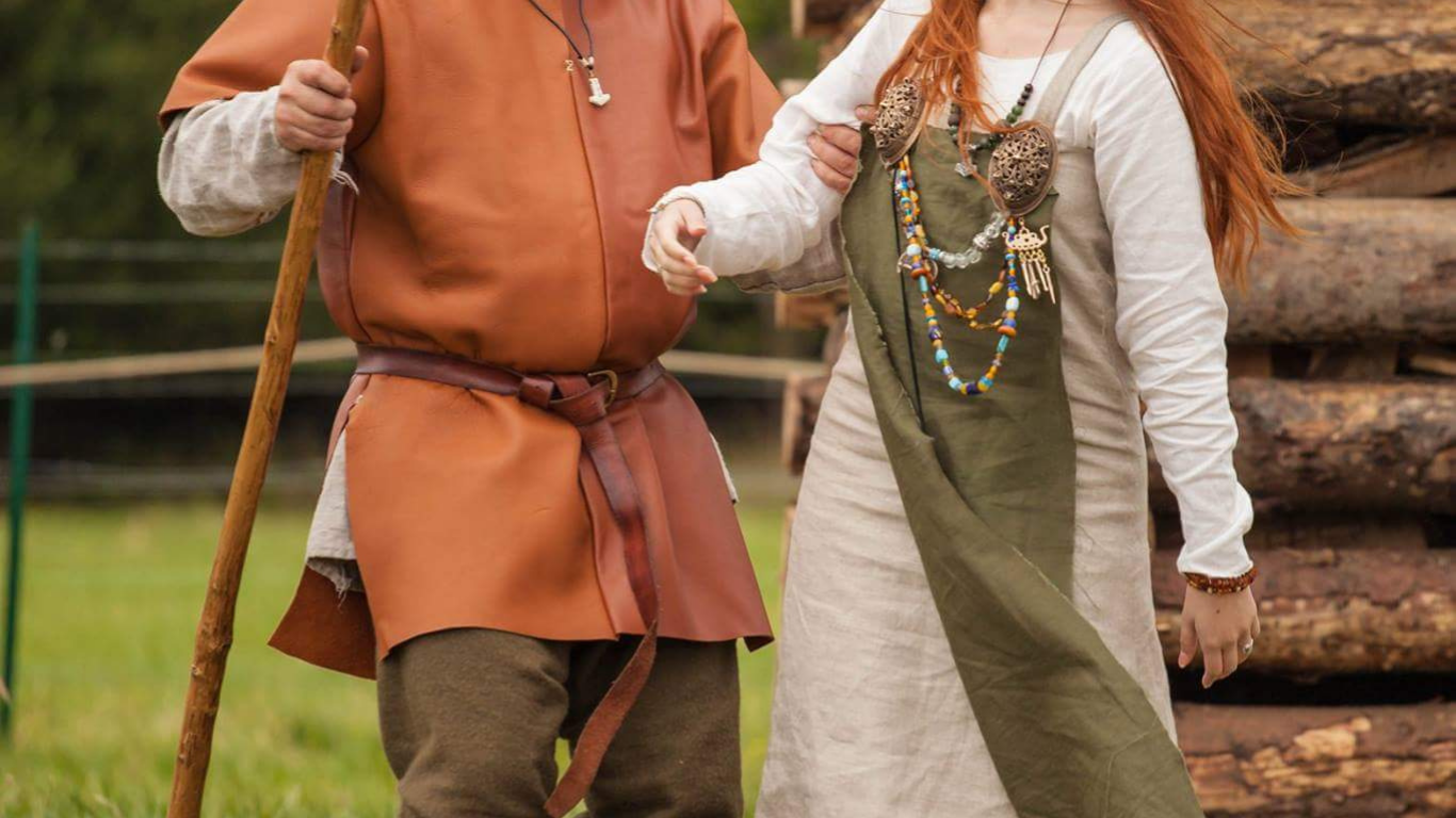 Deux personnes habillées de costumes médiévaux se tenant la main dans un extérieur avec des bûches en arrière-plan.