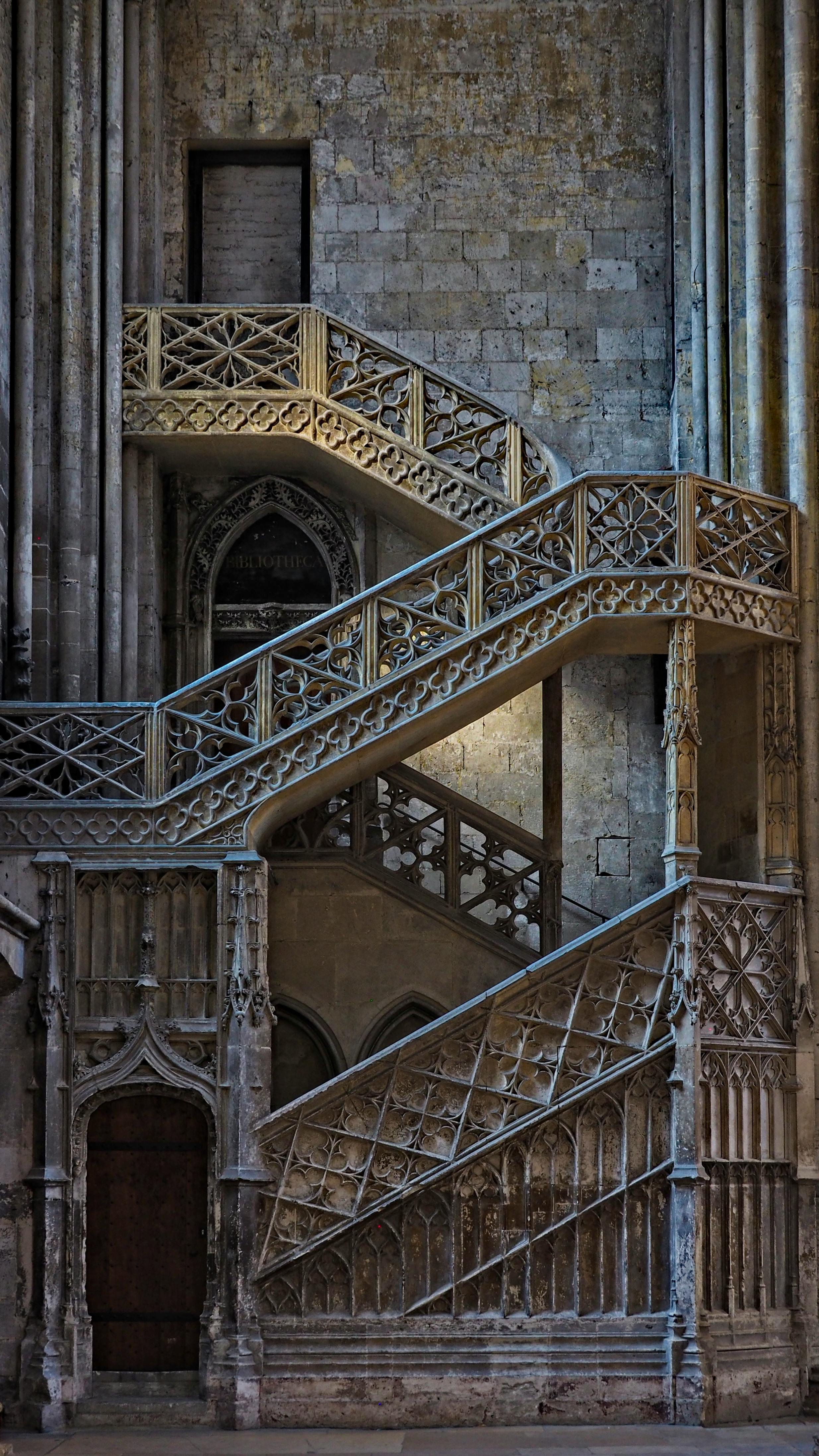 Escalier en pierre avec une balustrade en métal orné de motifs gothiques, intégré dans un mur en pierre ancienne.