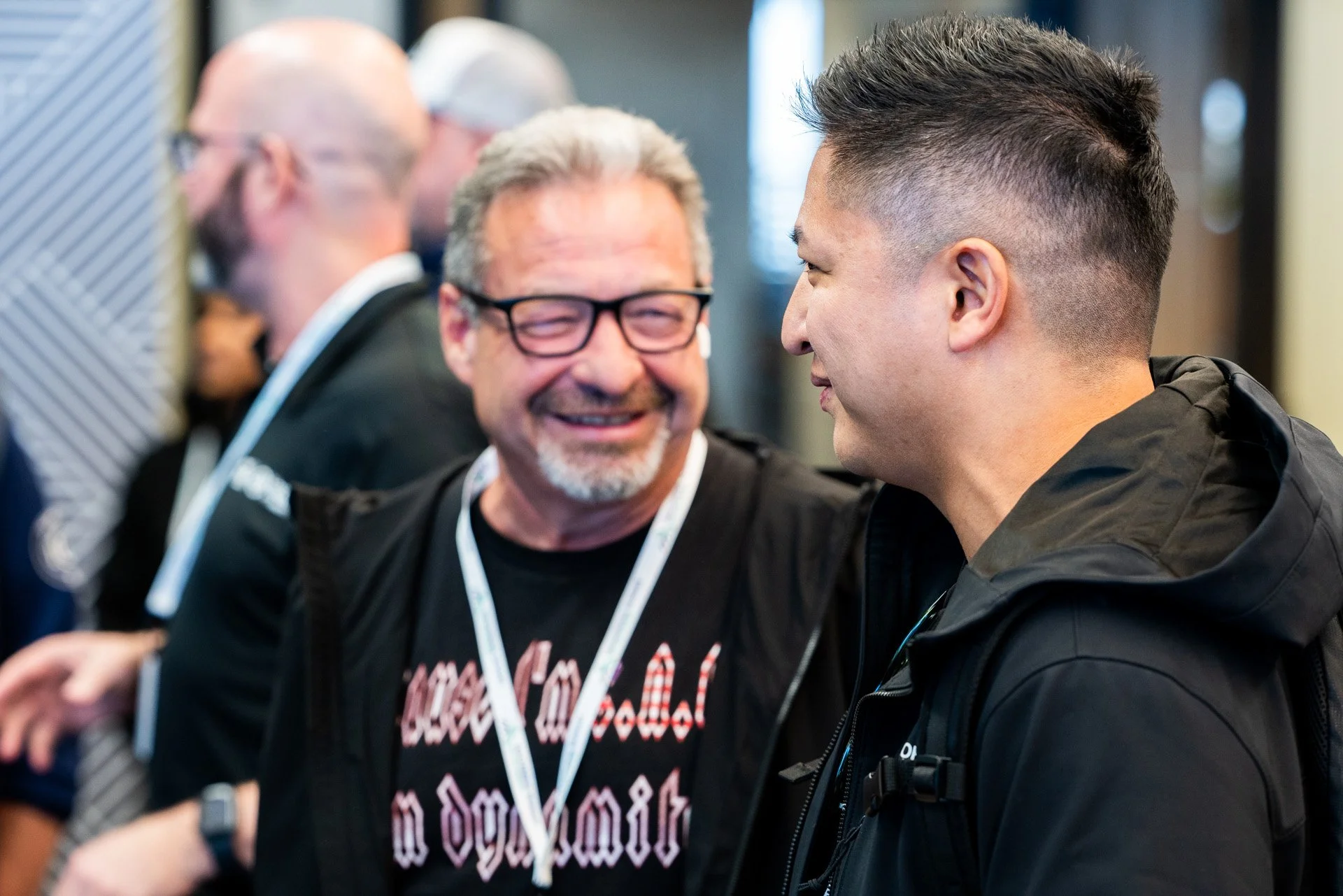 manufacturing leadership. Two men smiling and looking at each other, engaged in conversation, with a group of people in the background at an indoor event.