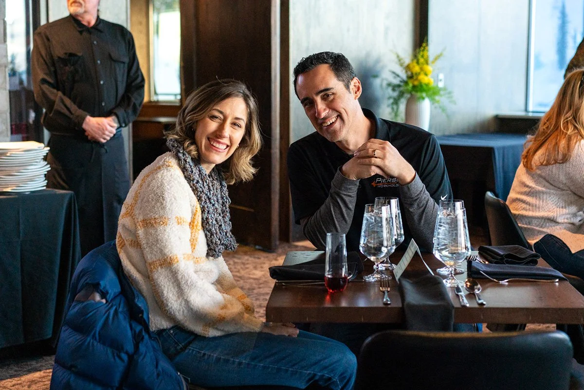 manufacturing industry community. A woman with shoulder-length hair and a scarf, and a man with short dark hair and a dark shirt, sitting at a restaurant table, smiling and enjoying dinner.
