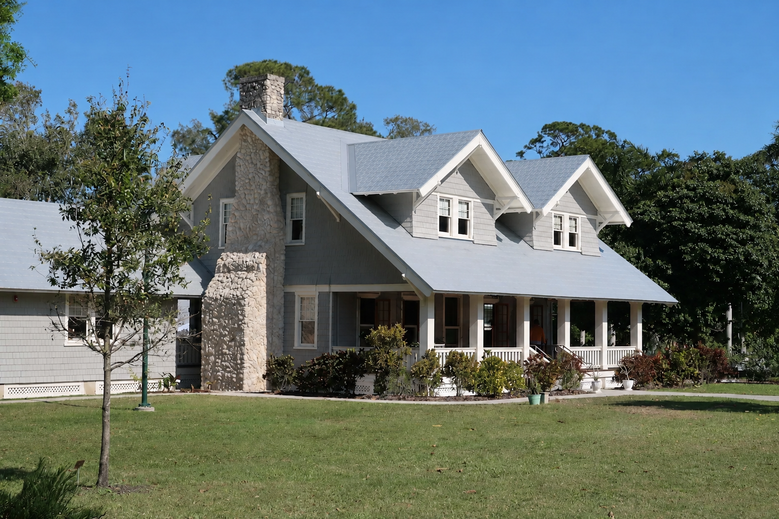 Gray residential home with a newly installed light-colored metal roof, stone chimney, covered front porch, and landscaped yard under a clear blue sky.
