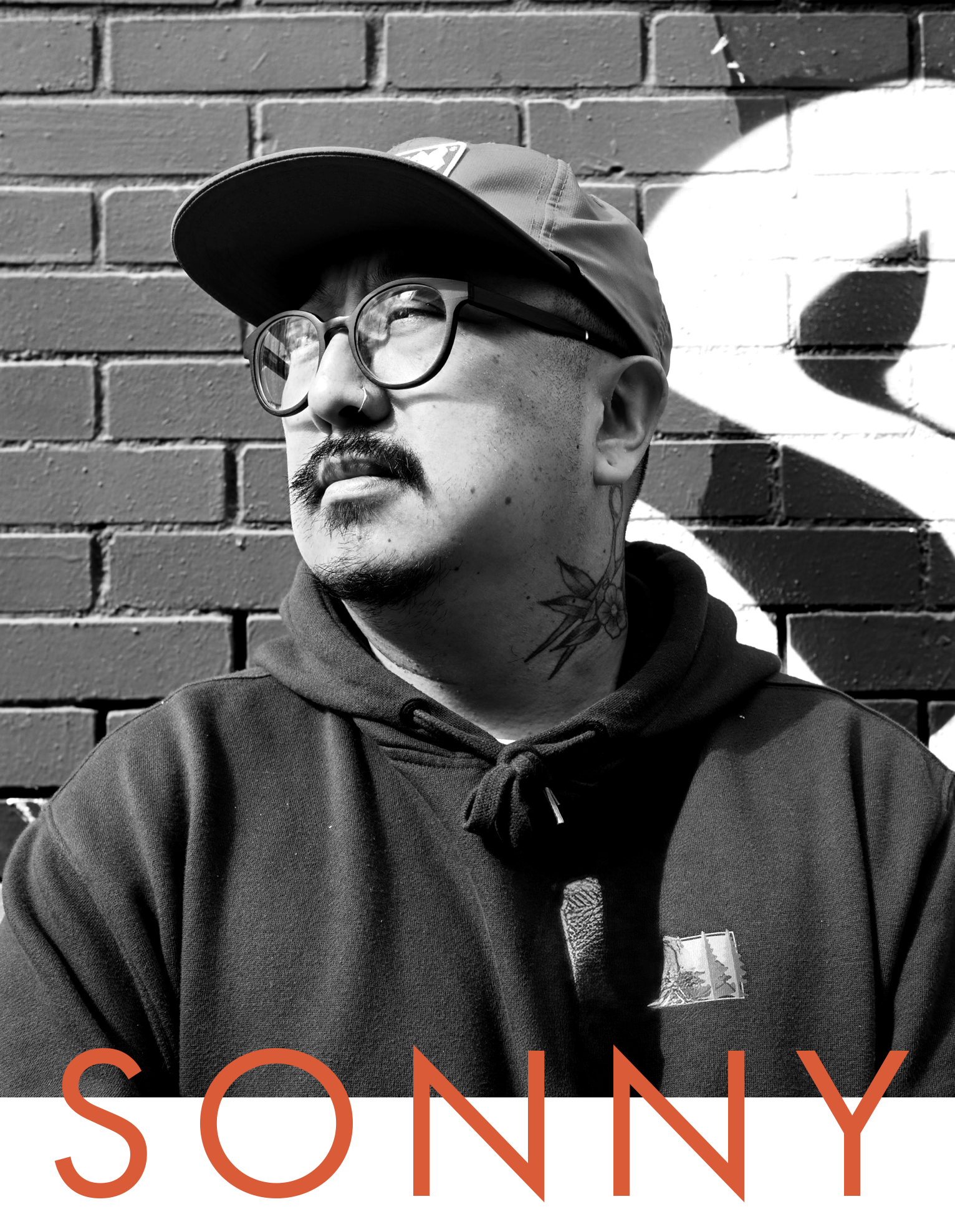 A black and white photo of a man with glasses, a mustache, and a tattoo on his neck, wearing a cap and hoodie, standing against a brick wall with painted graphics.