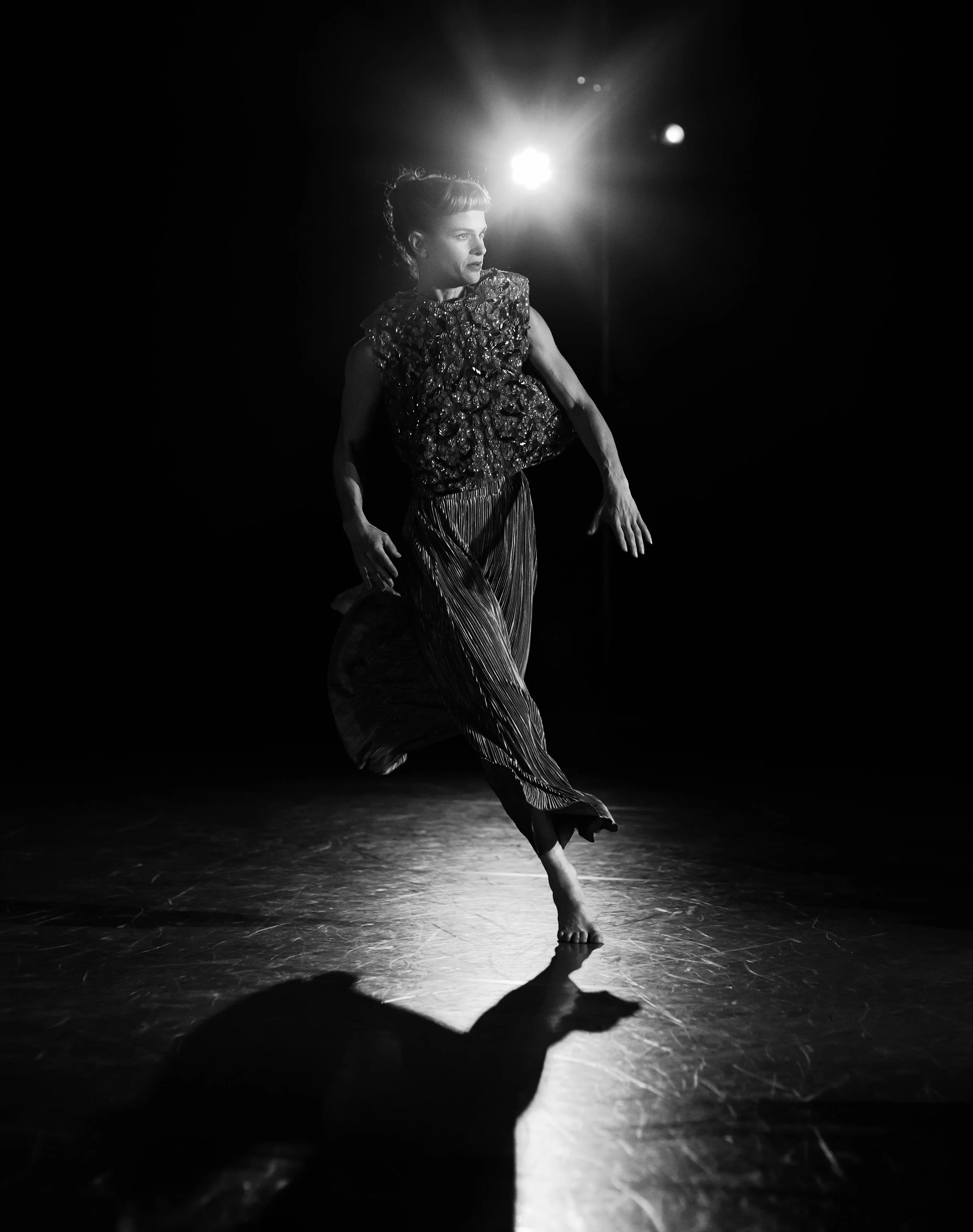 A female dancer performing on stage, illuminated from behind, wearing a textured top and flowing pants, with her hand extended and a shadow cast on the floor.