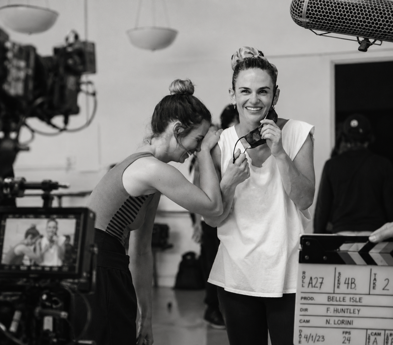Two women on a film set, laughing and enjoying a moment during production. One is holding a microphone, and there is a camera and a clapperboard in the scene.