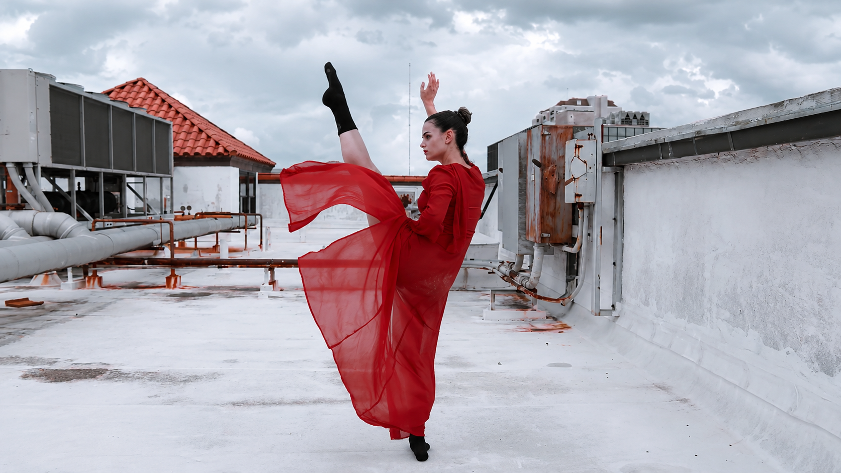 A woman in a red dress and black socks performs a ballet move on a rooftop with industrial pipes and equipment, under a cloudy sky.