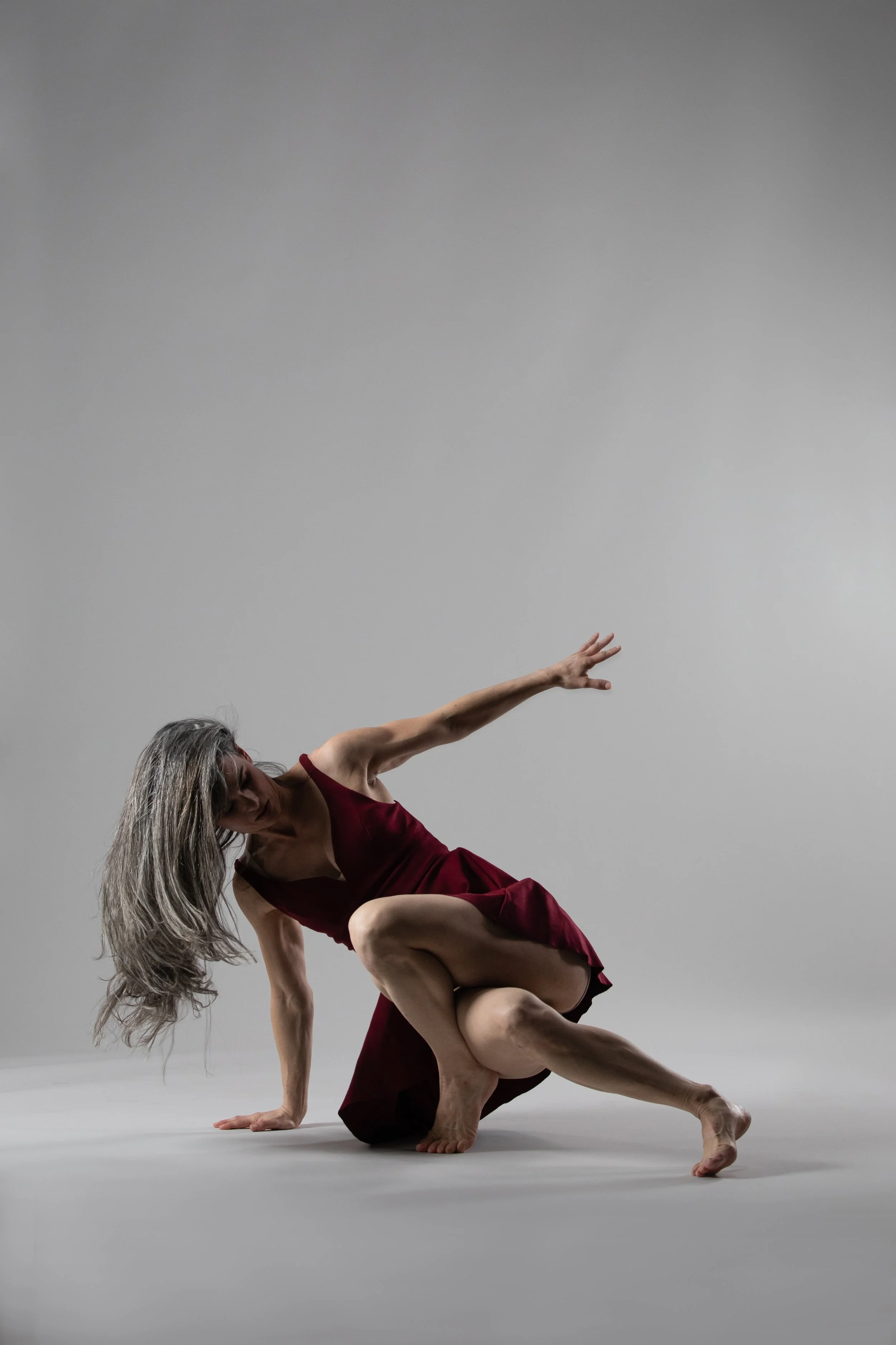 A woman with long gray hair in a red dress performing a contemporary dance move on a plain gray background.