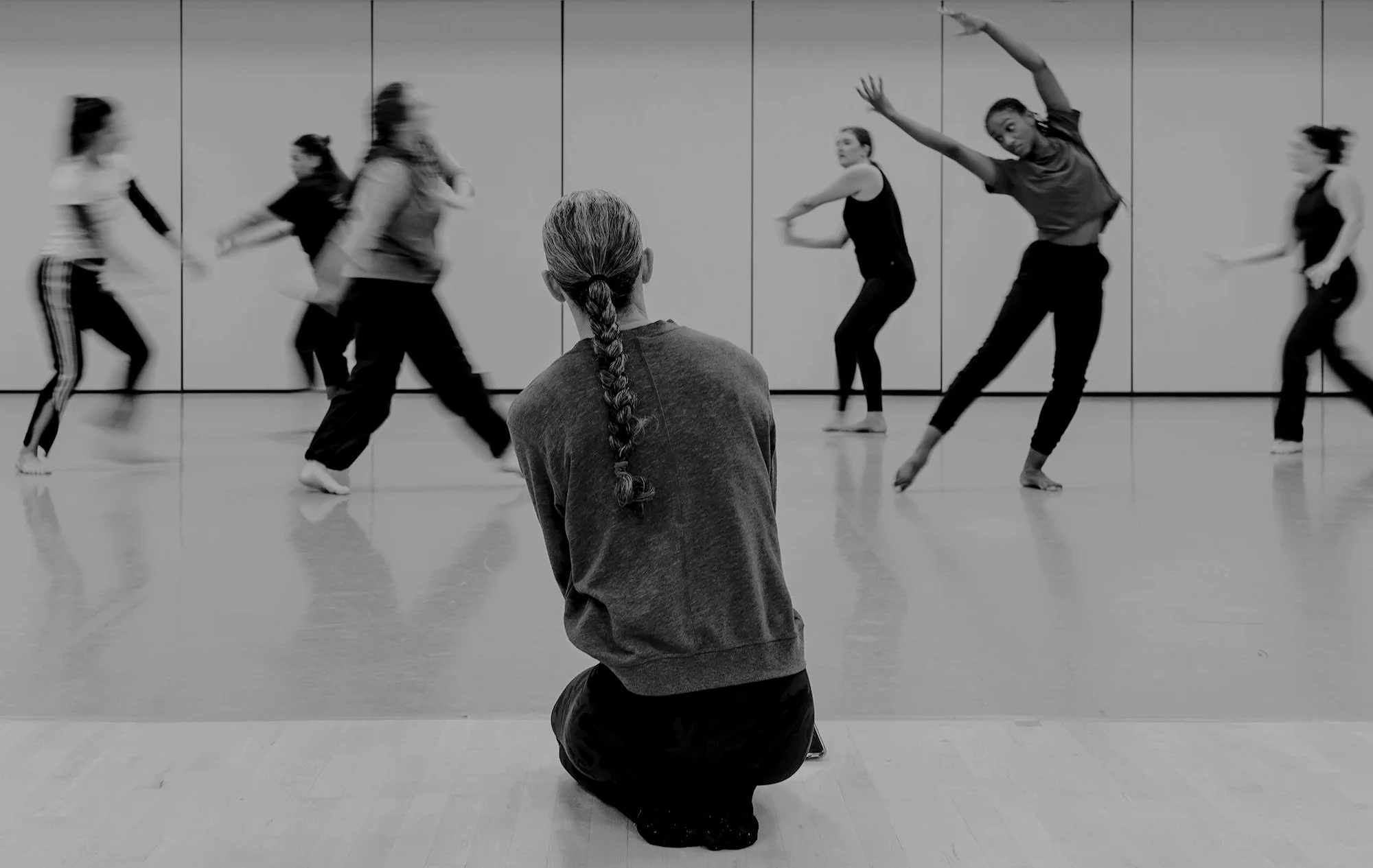 A person with gray hair in a braid, kneeling with arms crossed behind back, observing a dance class in a studio with a group of dancers practicing.