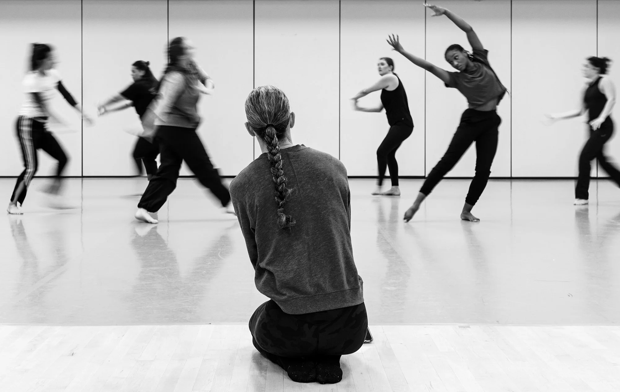 A person with braided hair sitting on the floor, watching a dance class with several women dancing in the background.