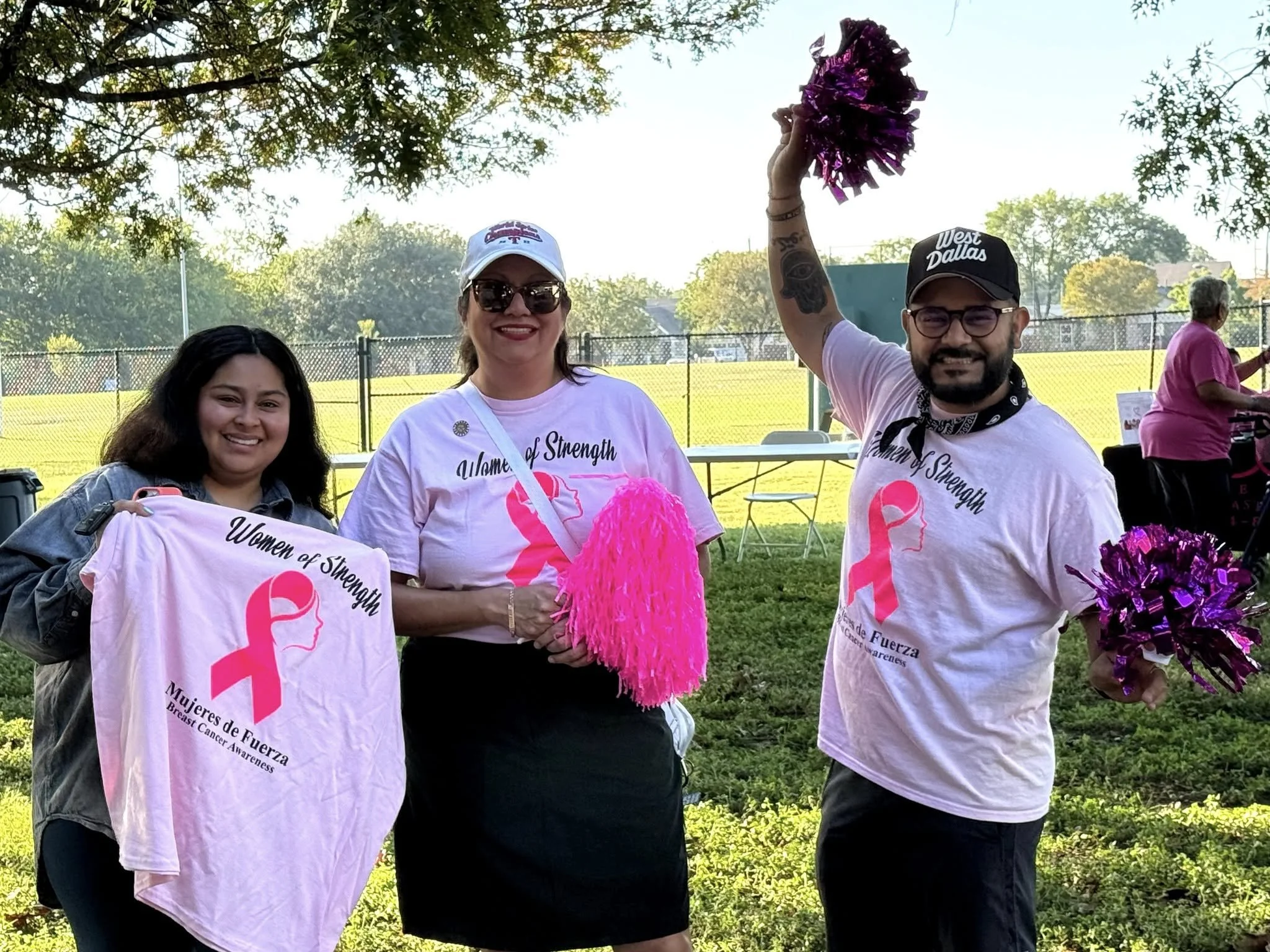 Group of three women smiling at an outdoor event, wearing pink Breast Cancer Awareness shirts, with one woman holding purple pom-poms and a cap that says 'West Dallas'.