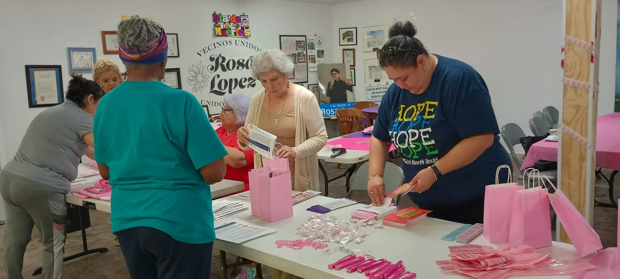 Several women are gathered around a table at an event, preparing pink-colored promotional materials and gift bags. The background features framed photos and certificates on the wall, with a sign that reads 'Vecinos Unidos Rosa Lopez.' The women are f