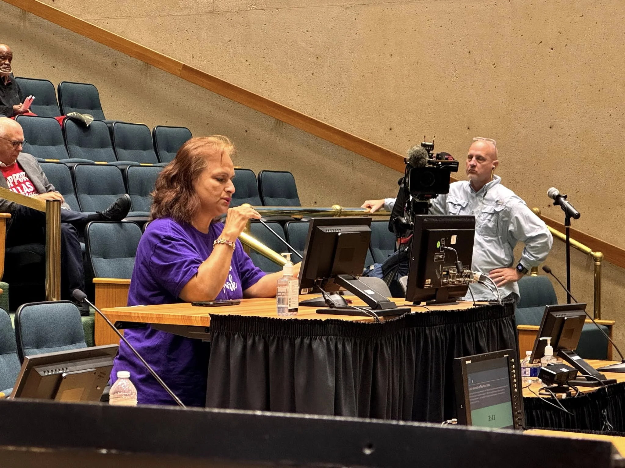 People sitting in a large conference room with tiered seating, some engaged with microphones, monitors, and cameras present.