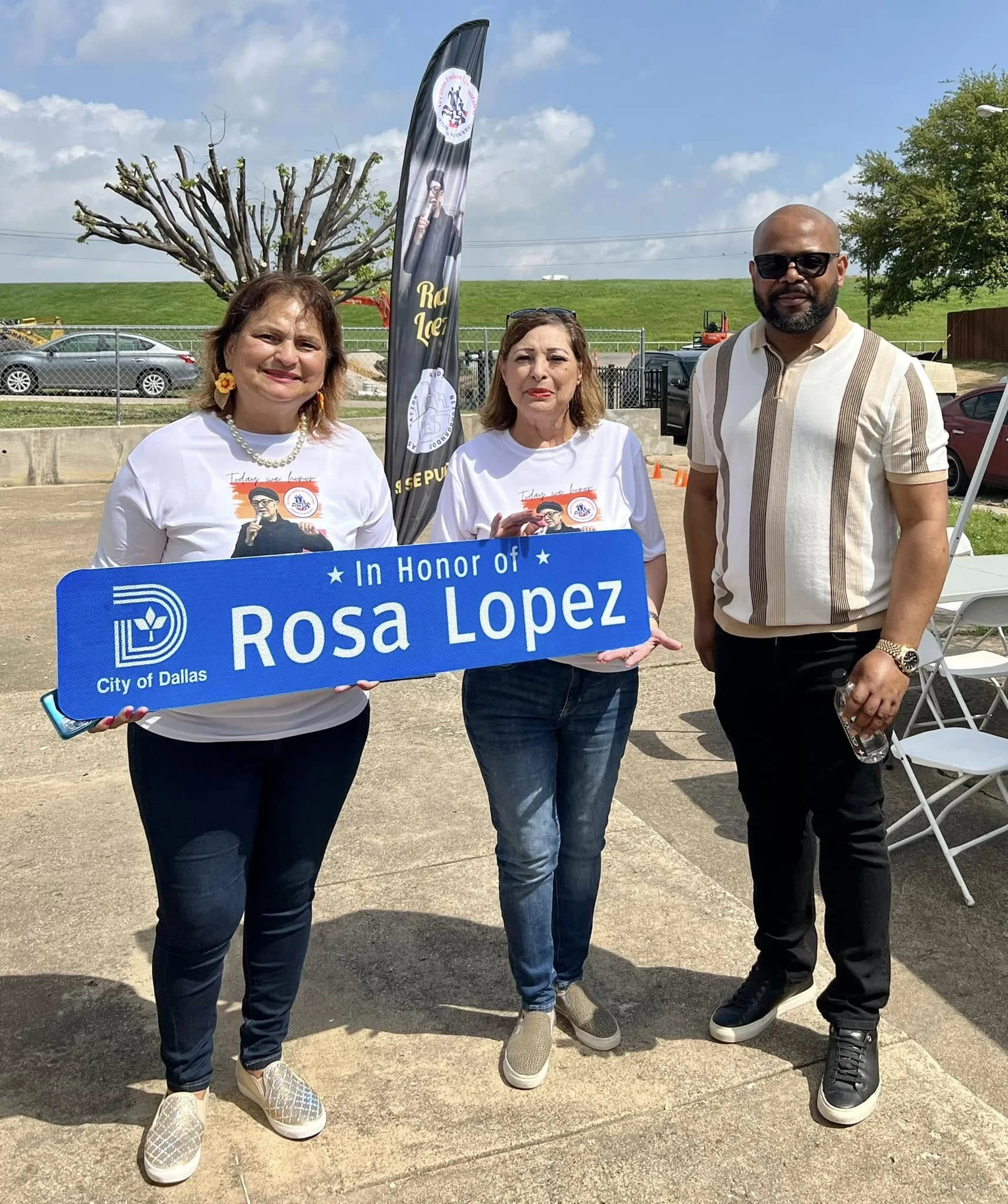Three people standing together outdoors, holding a blue sign that reads 'In Honor of Rosa Lopez' from the City of Dallas, with a black flag behind them. Two women are on the left, and a man is on the right. The sky is partly cloudy with some trees an