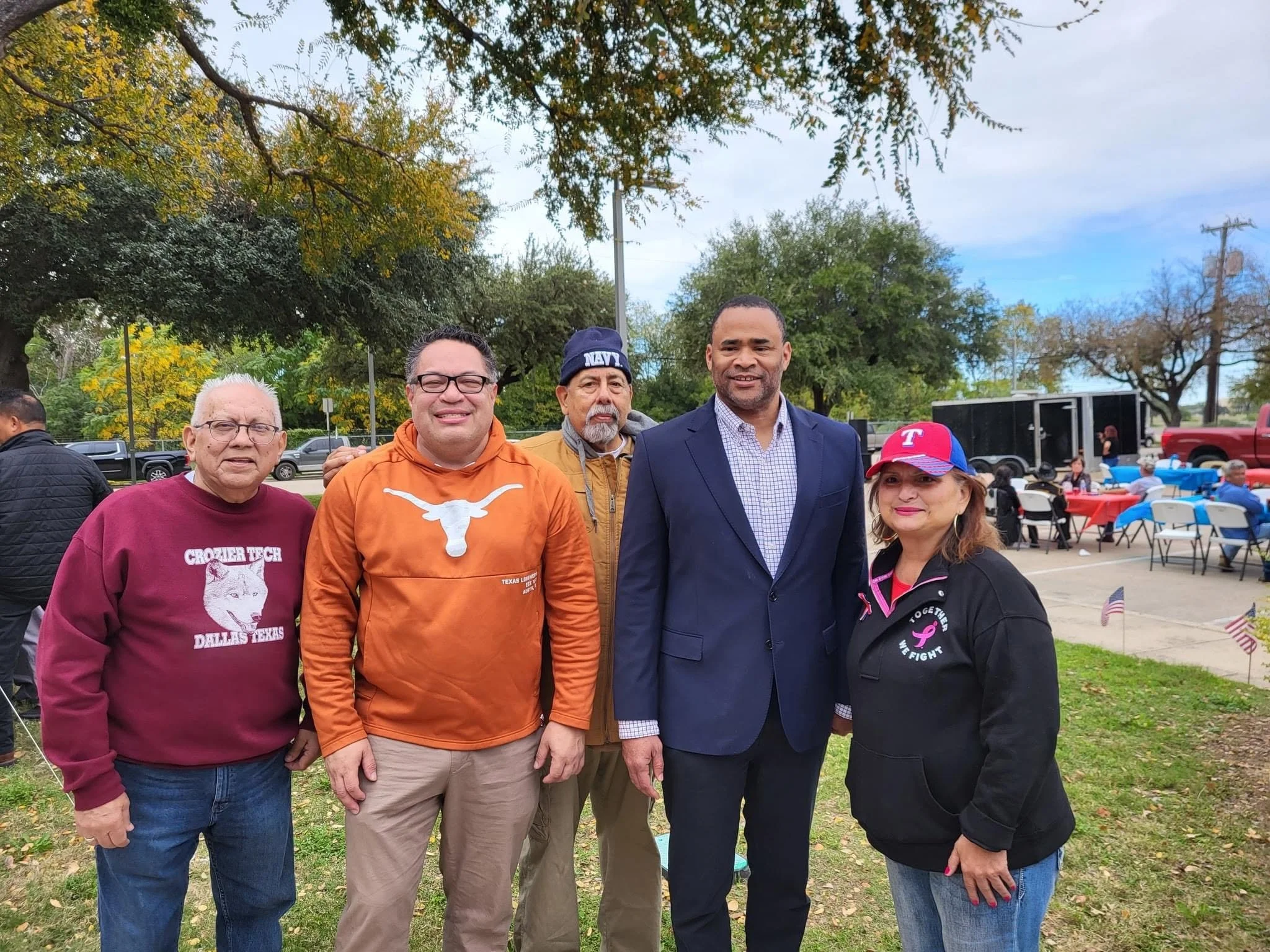 Group of five diverse people standing outdoors at a park during a community event, with trees, tents, and people sitting at tables in the background.
