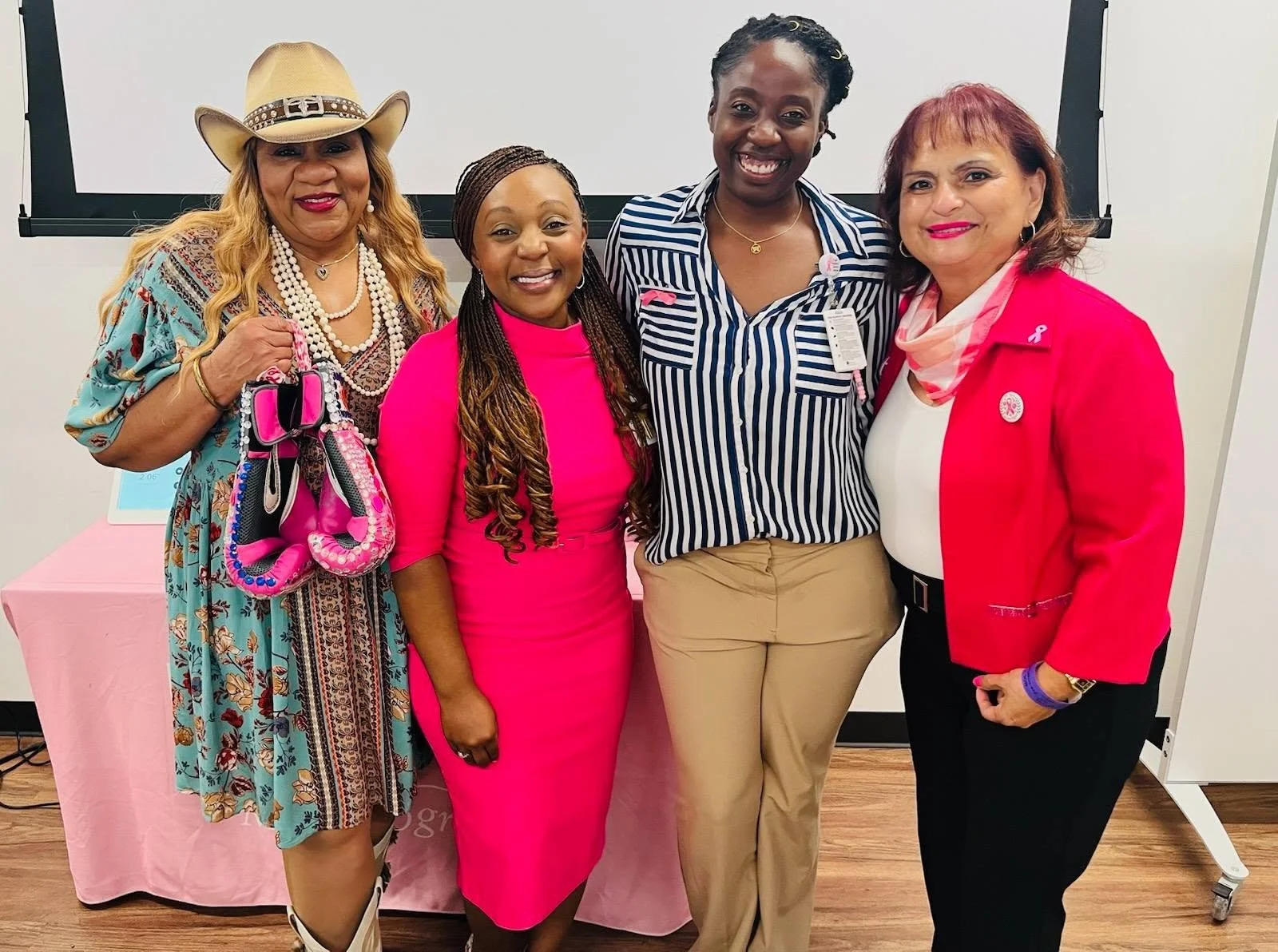 Four women standing together indoors, smiling at the camera. They are dressed in colorful clothing, with pink and red ribbons on their outfits, and a pink table with a laptop behind them.