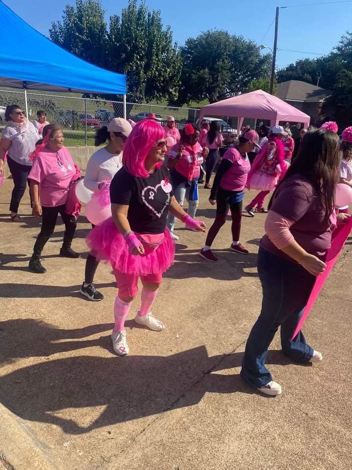 A group of women participating in an outdoor event, wearing pink-themed outfits and accessories, with a pink tutu, pink wigs, and pink balloons, in a community space with tents and trees.