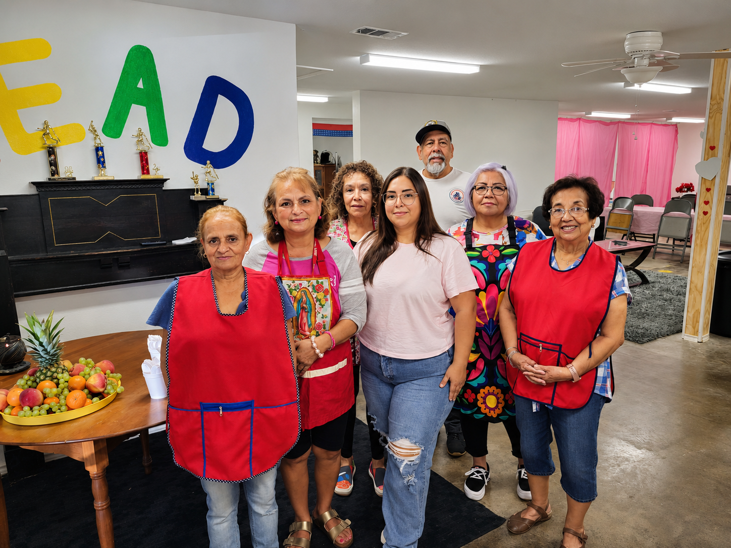 Group of seven women and one man standing in a community room decorated for a celebration, with 'FELIZ FAD' on the wall and a table of fruit.