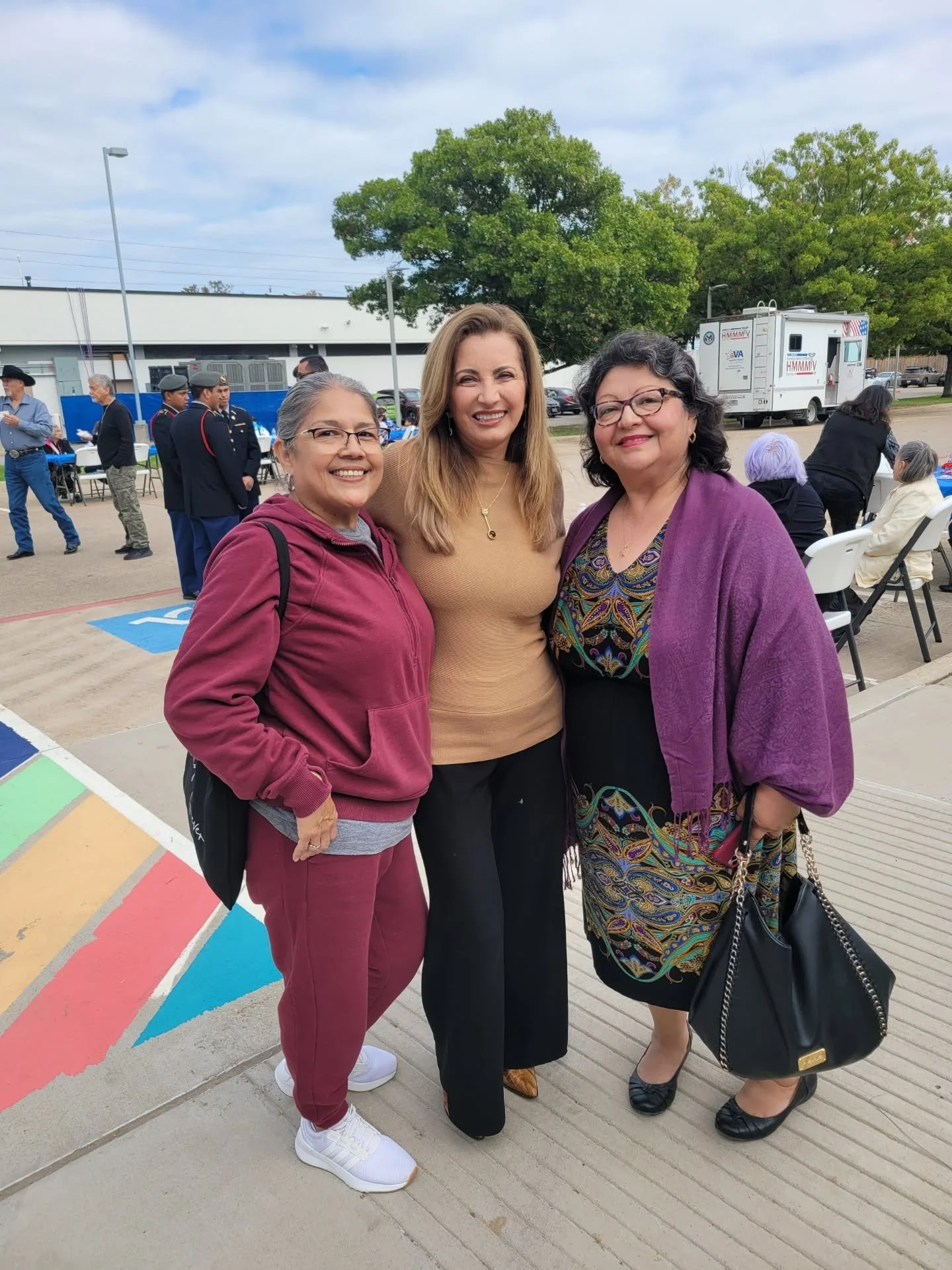 Three women standing together outdoors, smiling, with a group of people, trees, and a food truck in the background.