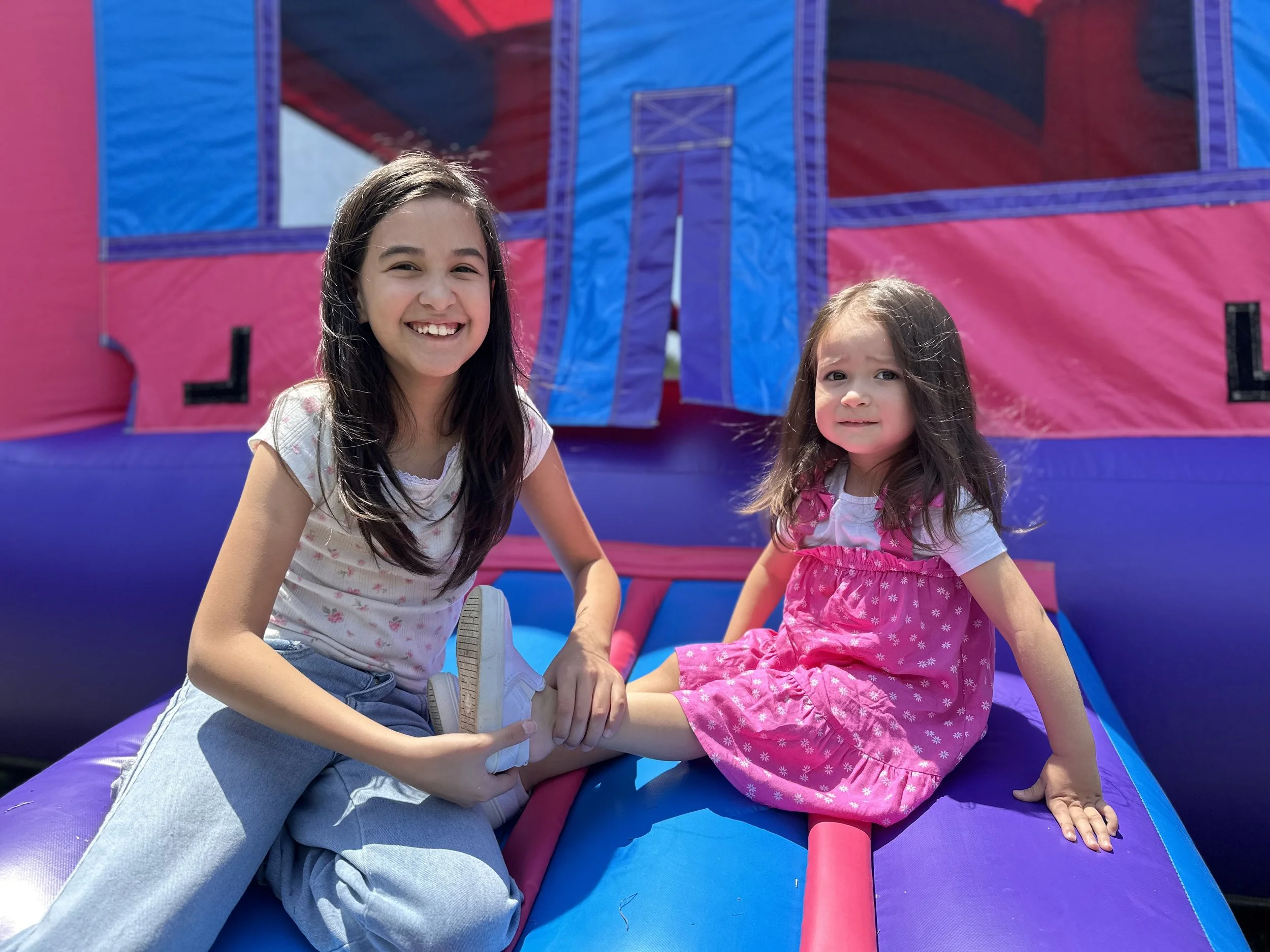 Two young girls sitting on an inflatable bounce house, one smiling and the other with a slightly concerned expression, with a pink and blue background.