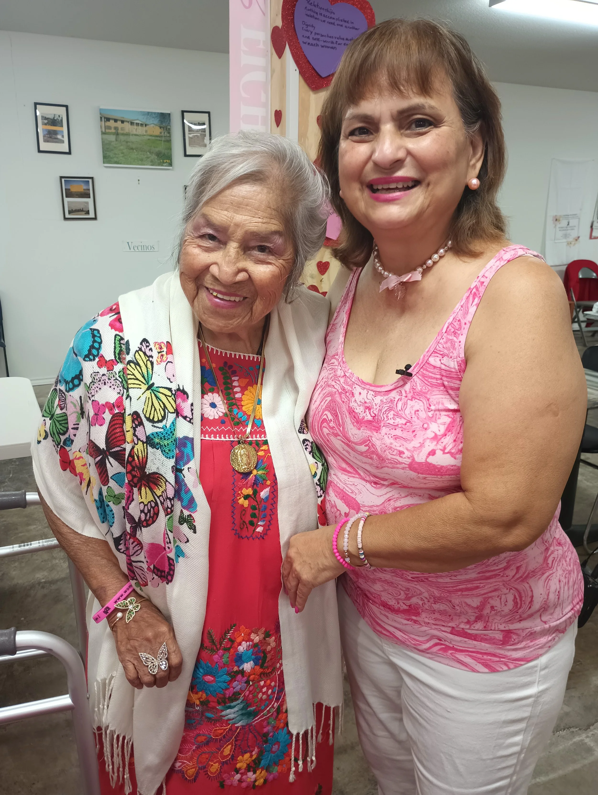 Two women smiling and posing for a photo indoors. The elderly woman on the left is wearing a colorful butterfly-patterned shawl and red dress with embroidered flowers, accessorized with a butterfly ring, a necklace with a pendant, and a bracelet. The middle-aged woman on the right is wearing a pink and white marbled sleeveless top, pearl earrings, a pearl necklace with a pink bow, and bracelets. The background includes framed photos on the wall and a sign that reads 'Vecinos'.