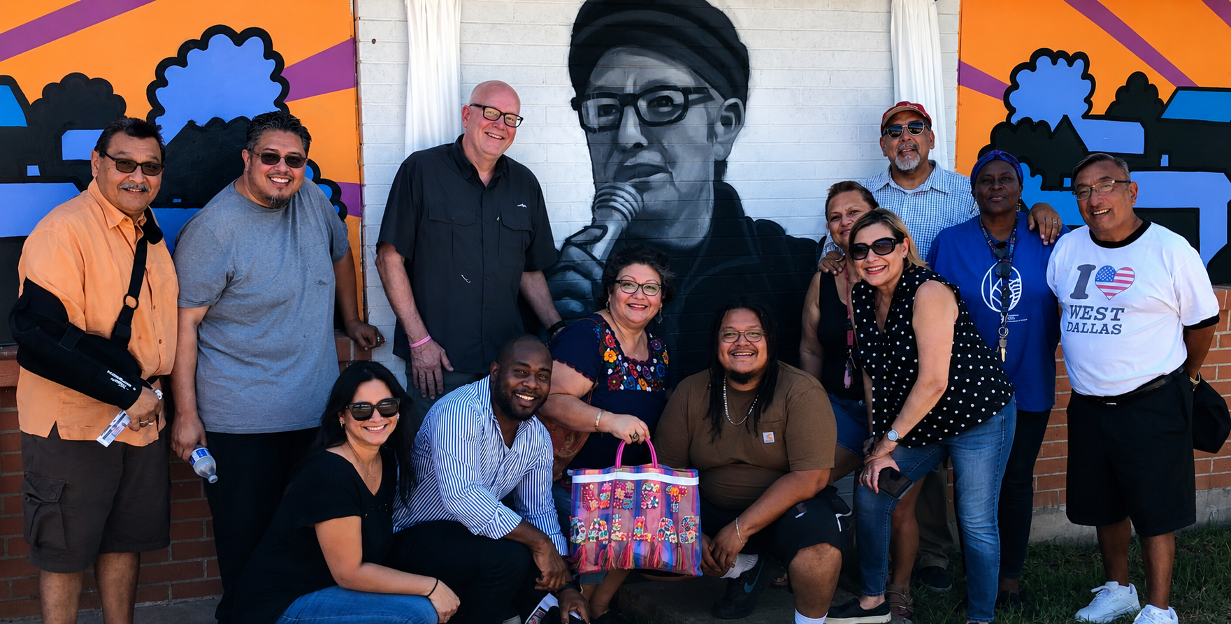A diverse group of people smiling and posing in front of a large mural of Roza Lopez n with glasses, holding a microphone. The mural is painted on a white brick wall with colorful background elements. Raul Reyes, is in this photo.