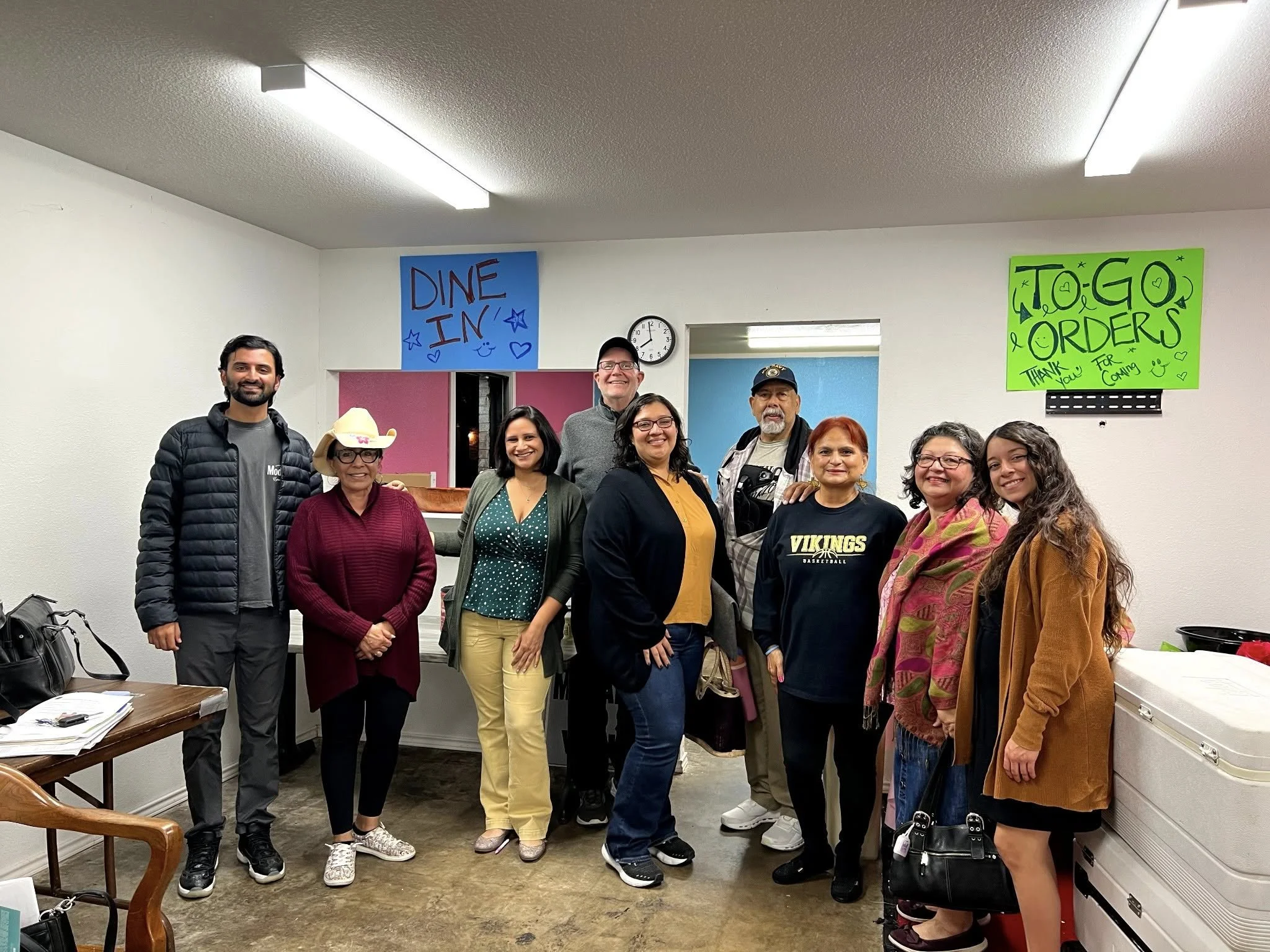 A group of ten people standing indoors, smiling at the camera, with signs on the walls that read "DINE IN" and "TO-GO ORDERS". The environment suggests a community or social gathering, possibly at a restaurant or community center.