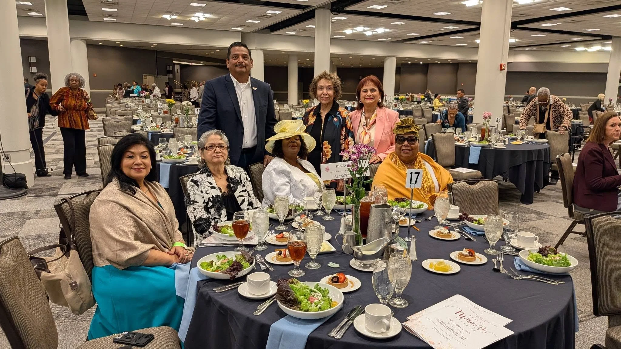 Group of women and one man seated and standing around a table at a formal event in a large banquet hall, with table settings including salads and drinks, and other guests in the background.