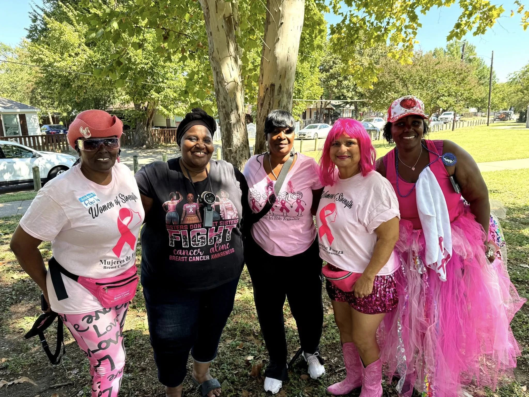 A group of five women standing outdoors in front of a tree, dressed in pink and black for breast cancer awareness. They are smiling, wearing pink shirts, accessories, and some have pink hair or pink tutu skirts. The background shows a sunny day, gree