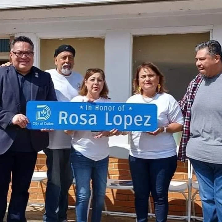 Group of five people holding a blue sign that reads 'In Honor of Rosa Lopez,' in front of a building with a brick wall and large windows.
