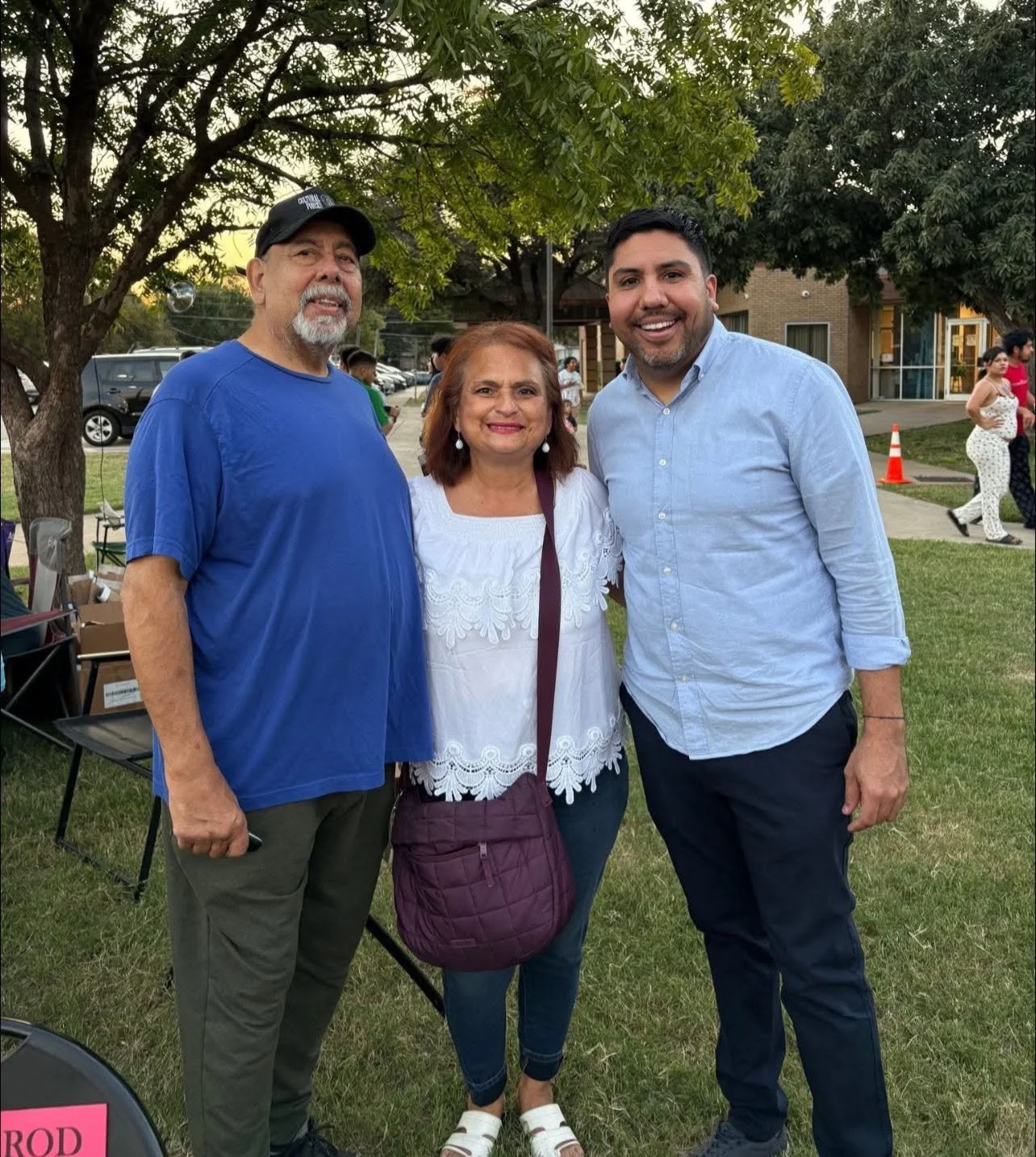 Three people stand outdoors on a grassy area, smiling at the camera. The man on the left wears a blue shirt and a black cap, the woman in the middle wears a white embroidered blouse and has a maroon purse, the man on the right wears a light blue butt