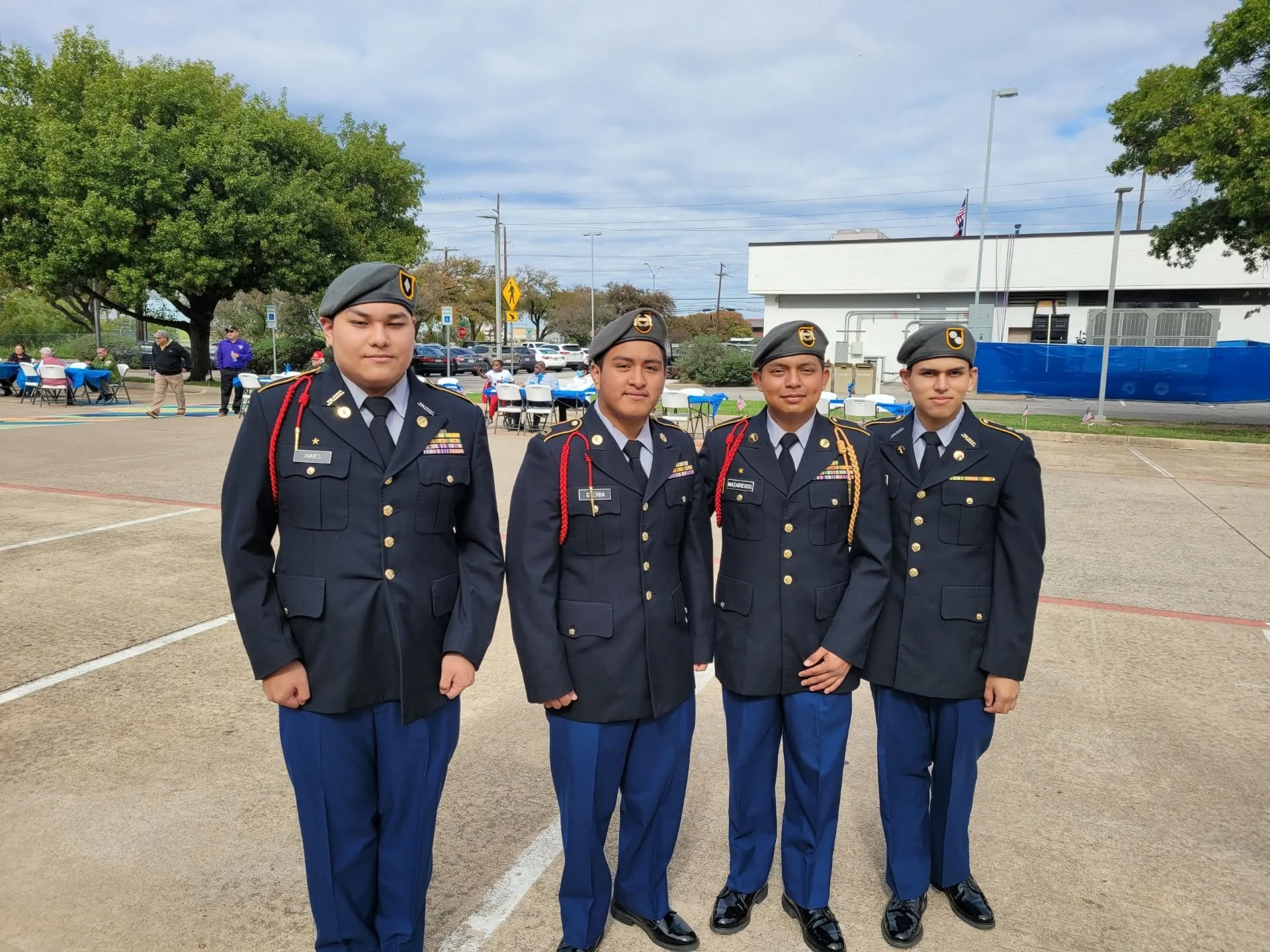 Four young men in military dress uniforms standing outdoors. They are in a formation, and there are tables and people seated in the background at an event or gathering.