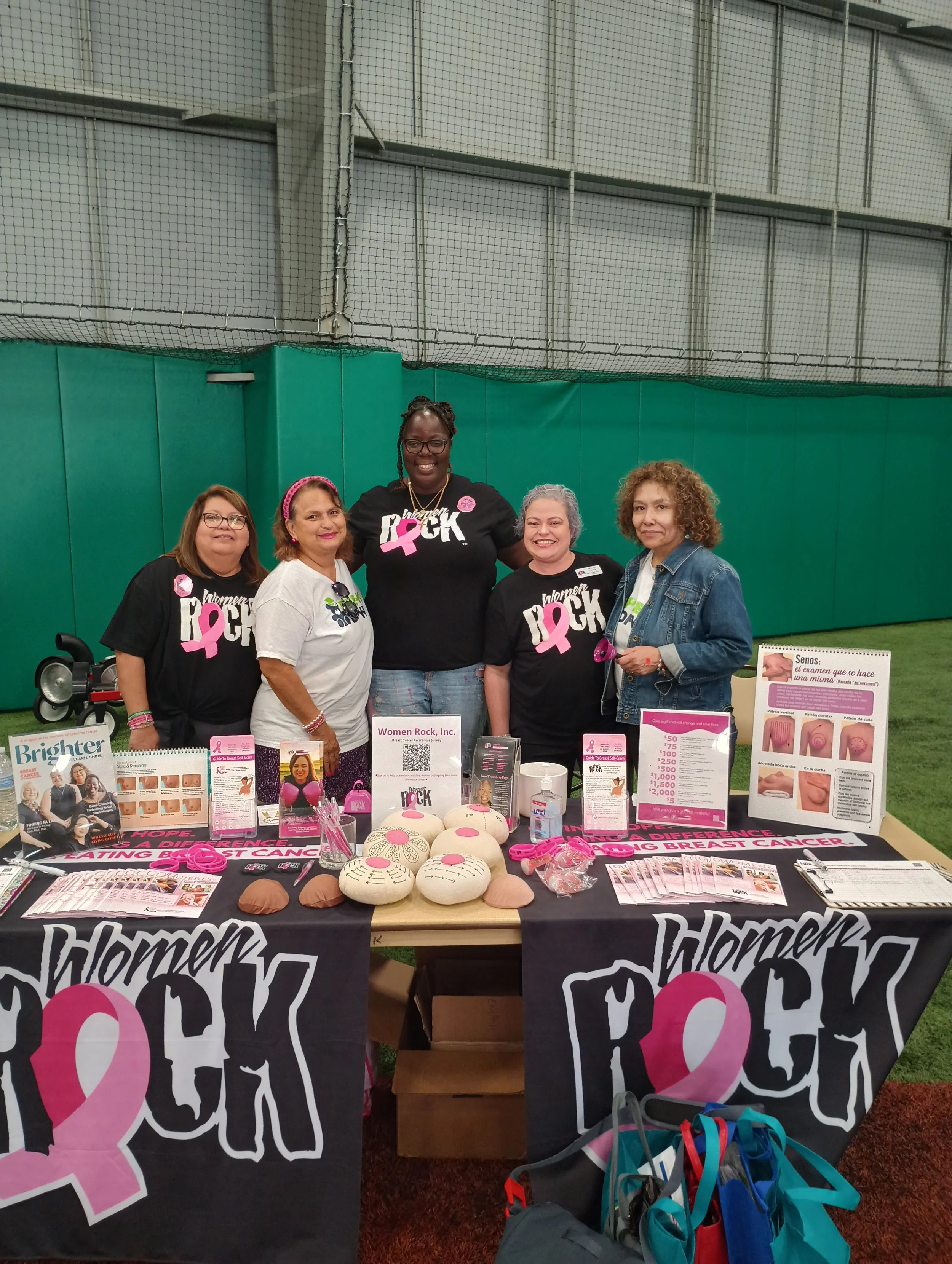 A group of women standing behind a table with breast cancer awareness materials, plush breast models, and informational posters, inside a sports facility.