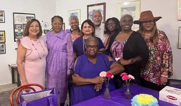 Group of eight women gathered around a table with birthday cake and decorations, celebrating together in a room with framed pictures on the wall.