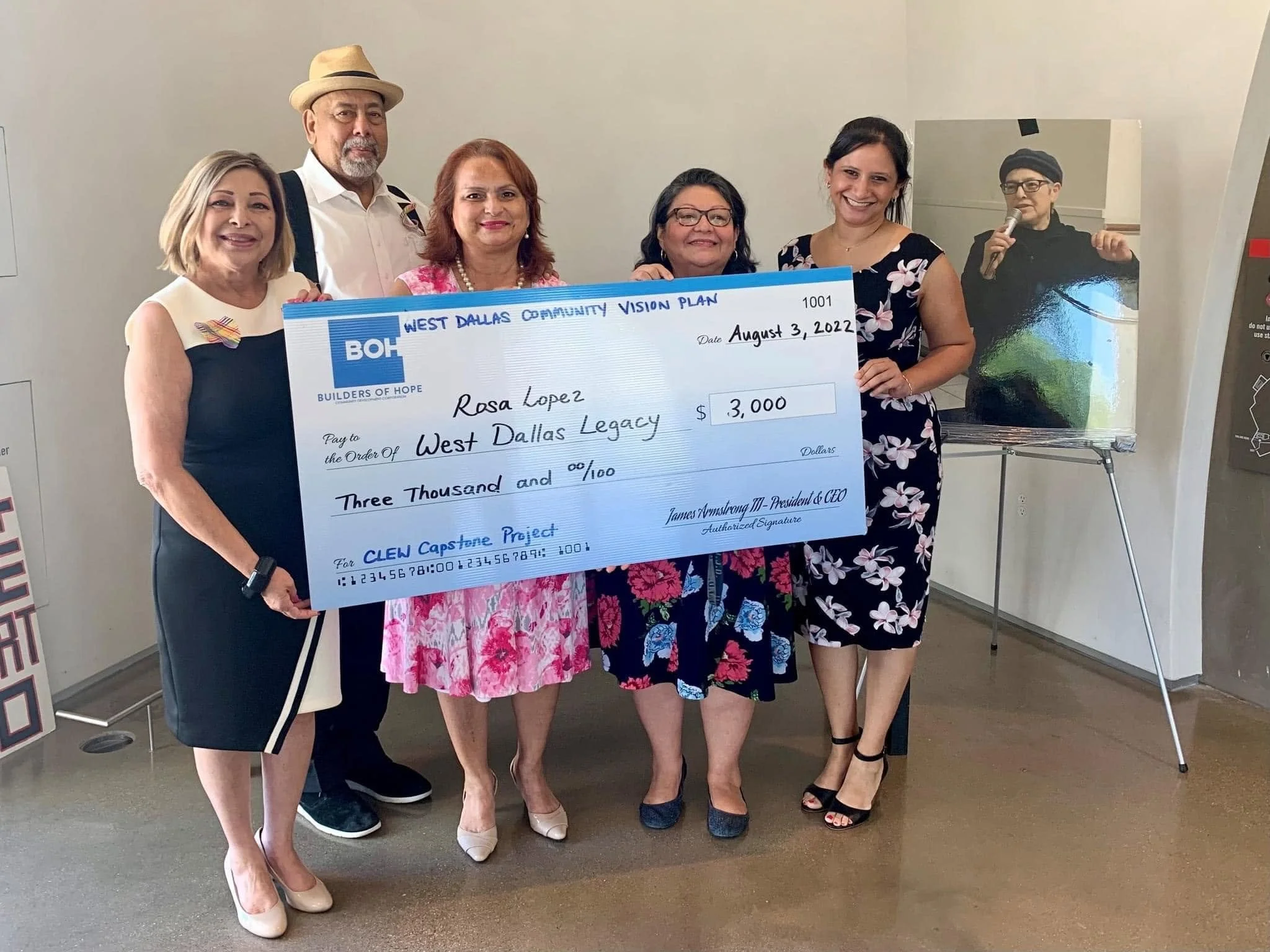 Group of six women and one man standing indoors, holding a large check for $3,000 made out to West Dallas Legacy, celebrating a community project, with posters and an image of a woman speaking into a microphone in the background.