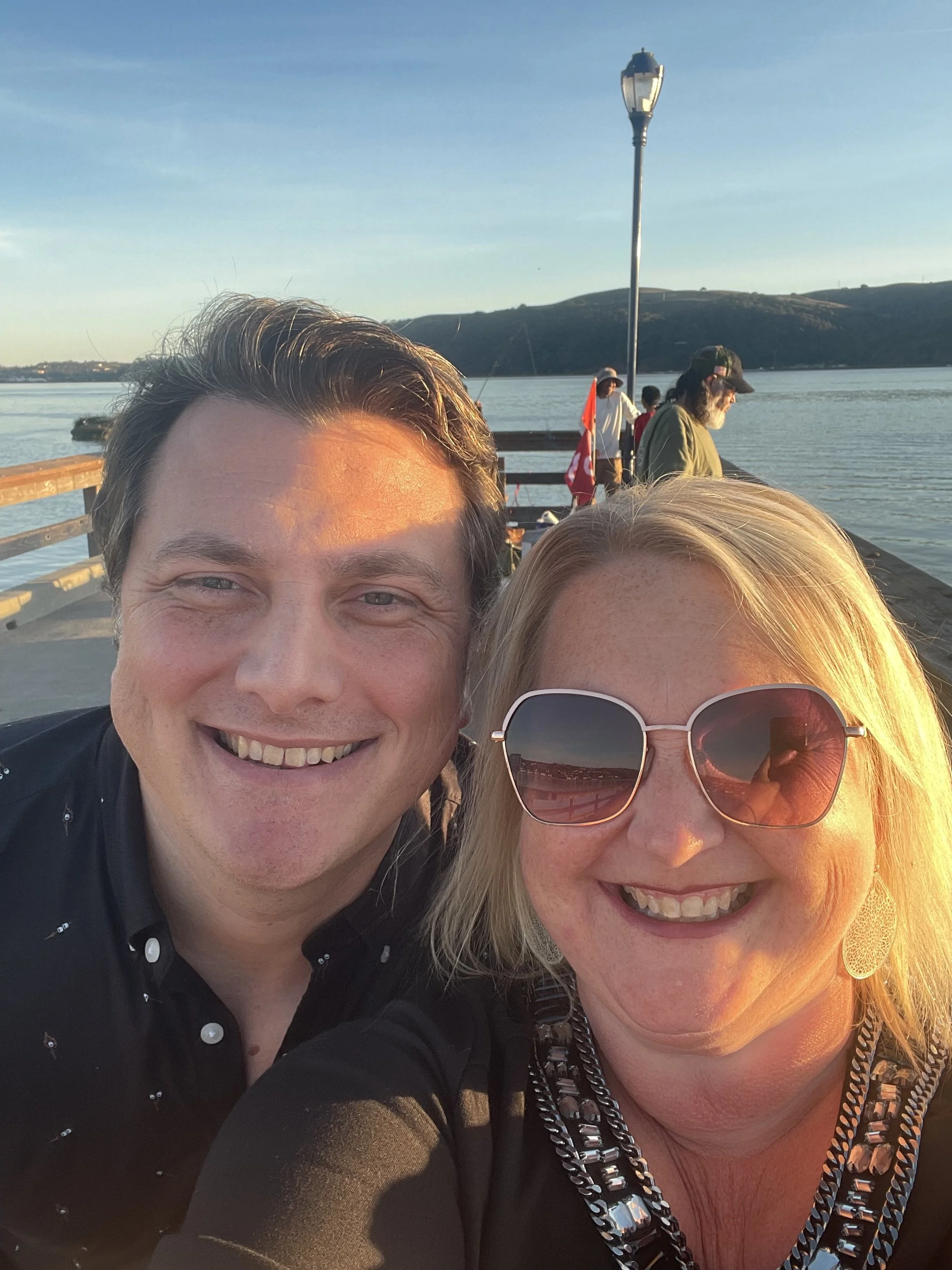 Close-up of a smiling man and woman taking a selfie outdoors by a body of water with hills in the background, during sunset.