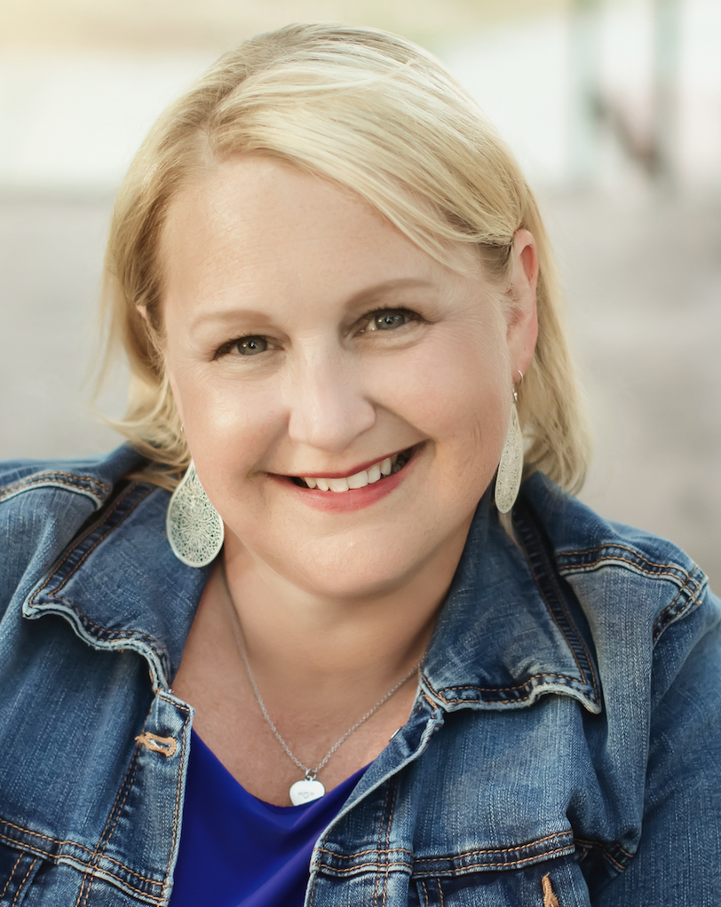 A smiling woman with blonde hair wearing a denim jacket, earrings, and a necklace, sitting indoors with a blurred background.