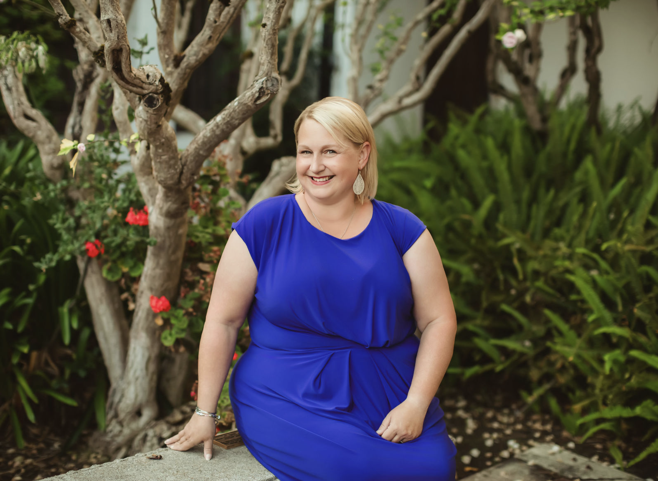 A woman in a royal blue dress sitting outdoors near a tree with green foliage and pink flowers, smiling at the camera.
