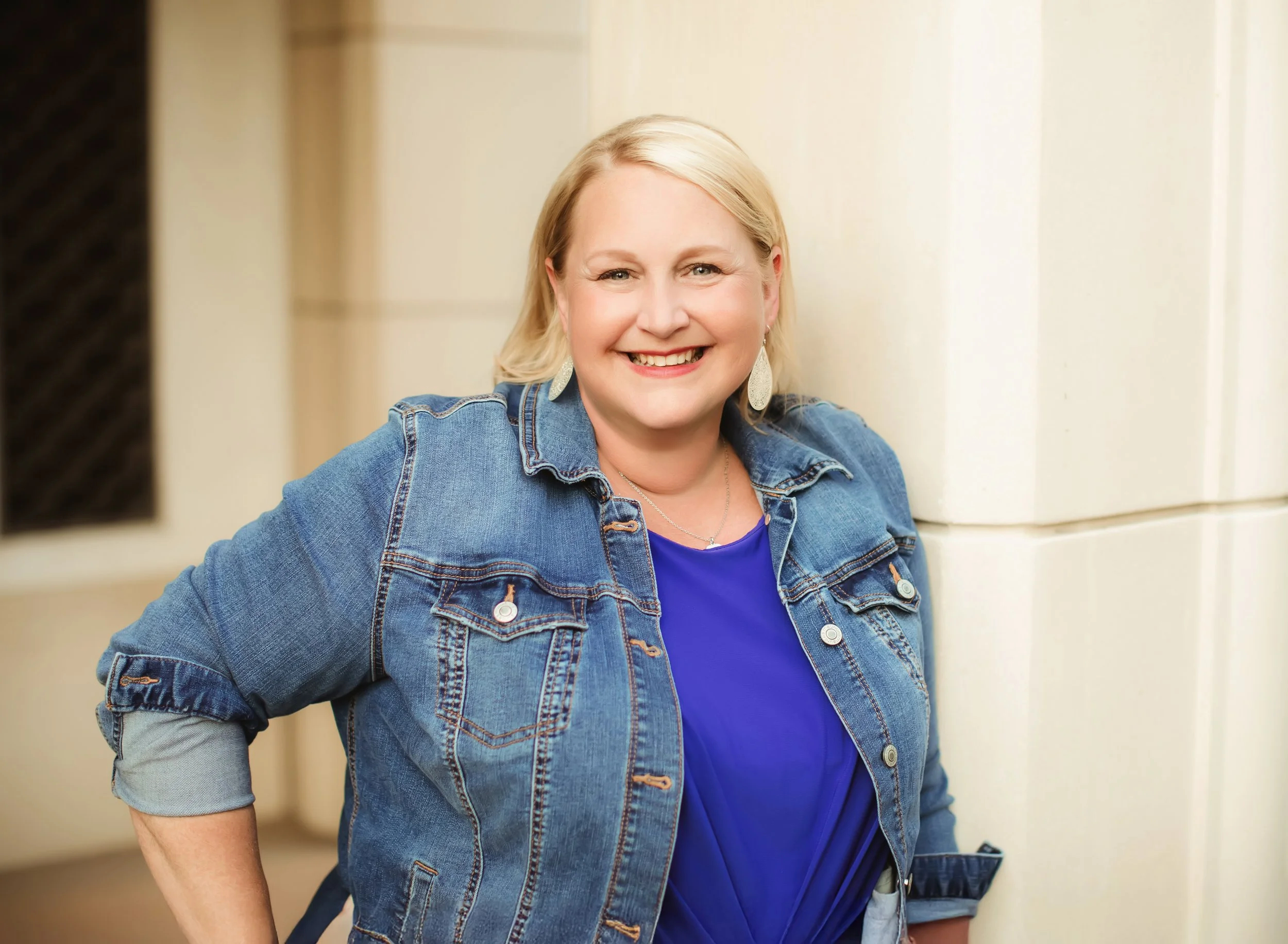 A smiling woman with blonde hair, wearing earrings and a denim jacket over a blue top, standing outdoors near a light-colored building.
