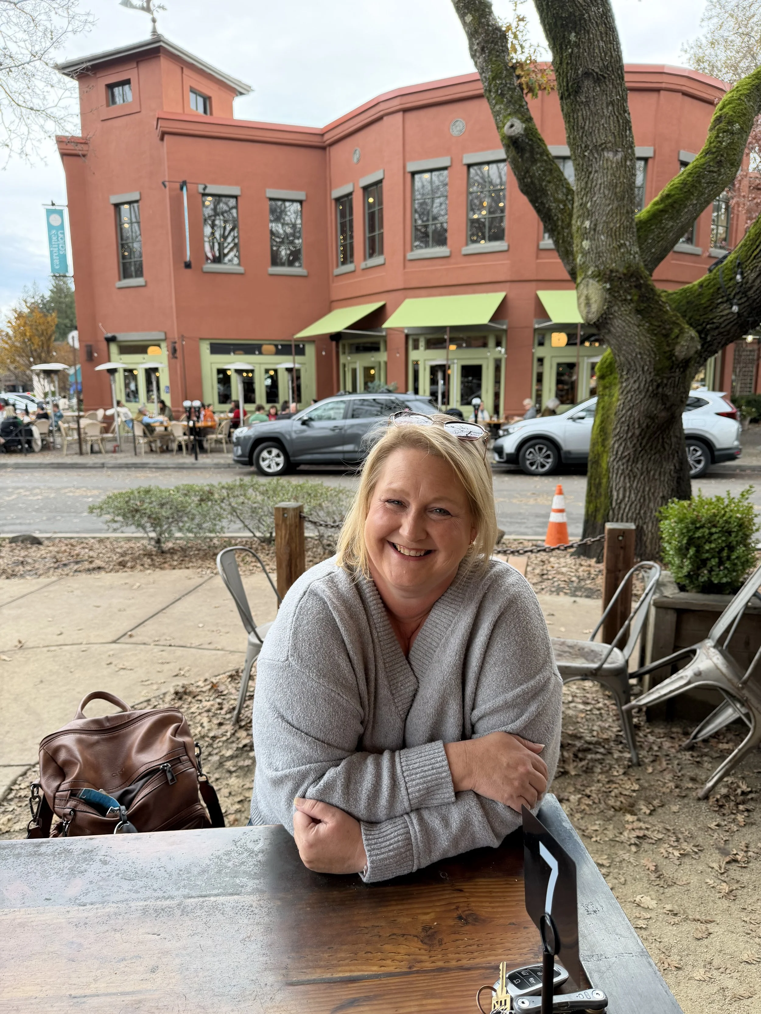 A woman sitting at an outdoor cafe table with her arms crossed, smiling. In the background, there are other people dining outside, parked cars, and a red brick building with green awnings and several windows. A large tree is in the foreground.