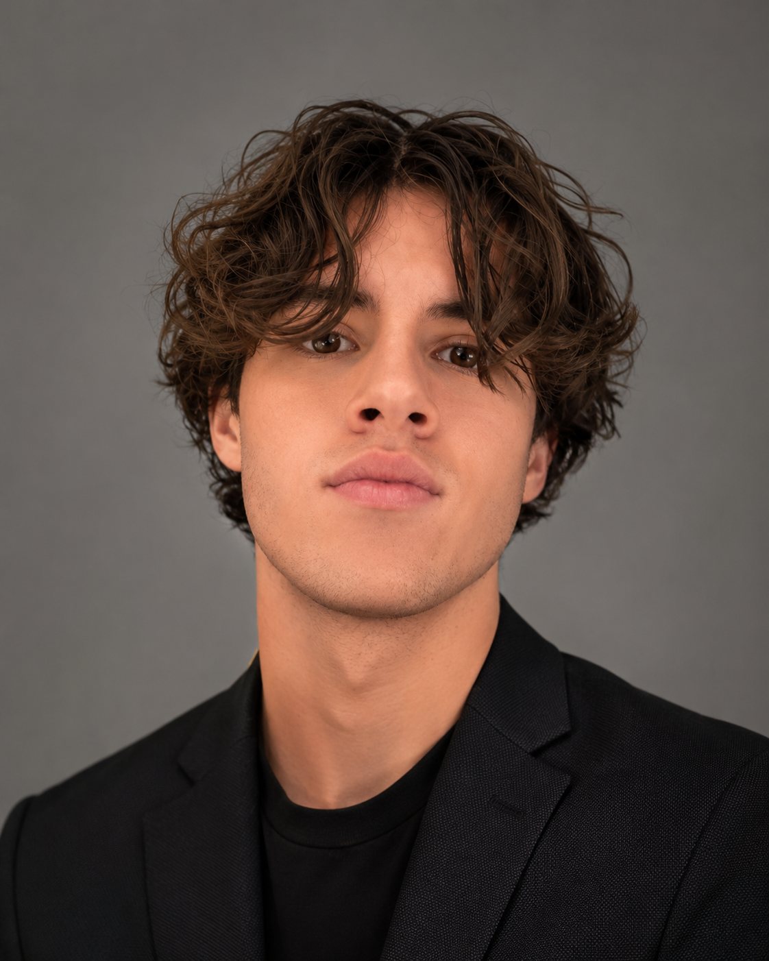 Close-up portrait of a young man with wavy brown hair, light skin, wearing a black blazer and T-shirt against a gray background.