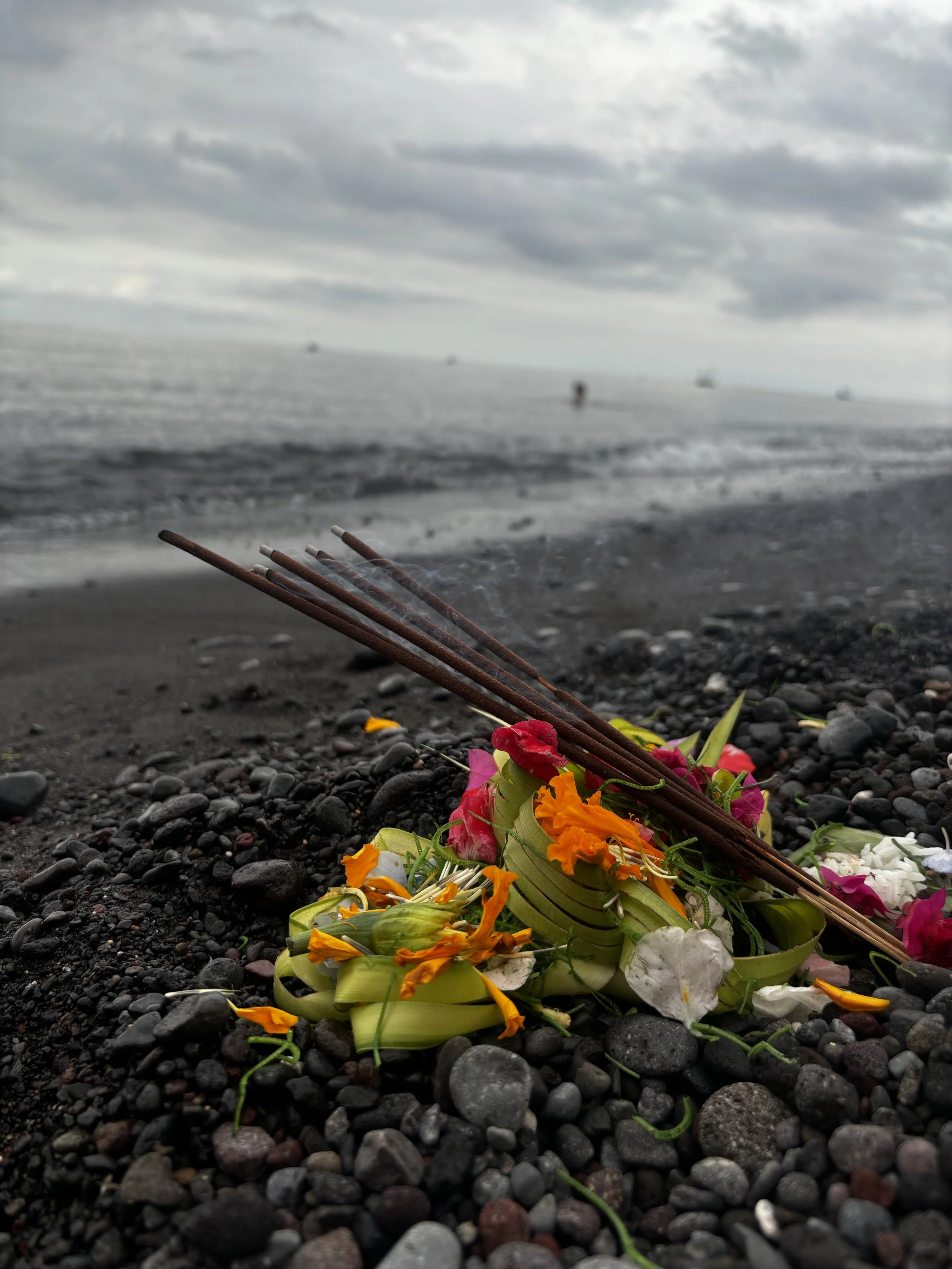 A small floral arrangement of colorful flowers, incense sticks, and a green banana leaf, lying on a pebble beach near a cloudy ocean, with boats visible on the horizon.