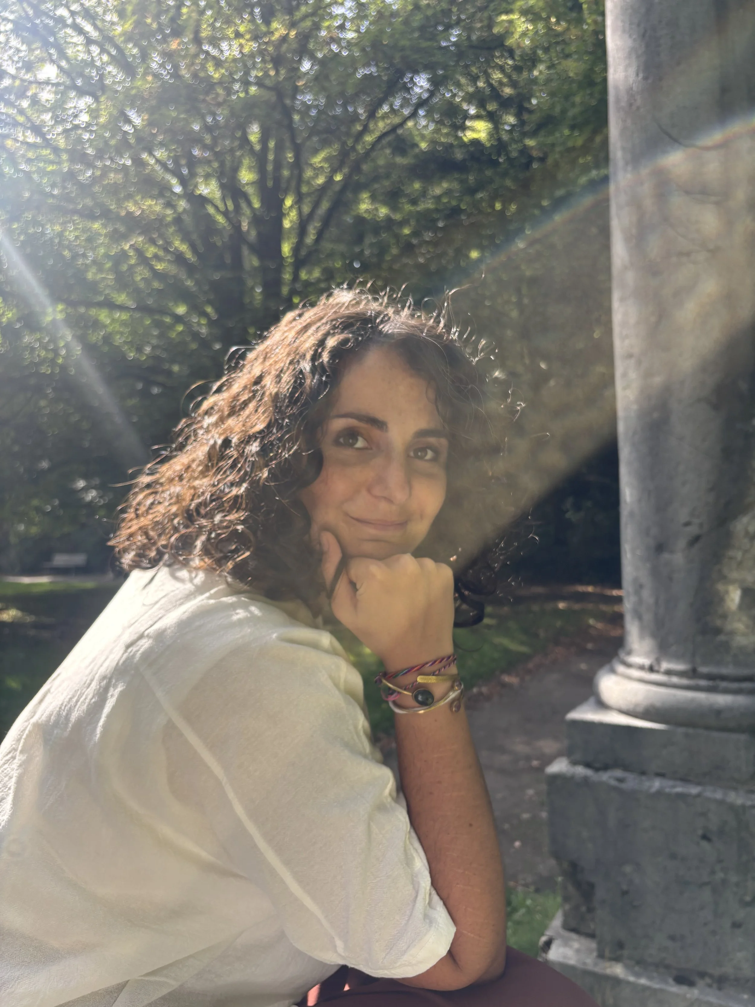 A woman with curly brown hair and light skin smiling, resting her chin on her hand. She is outdoors during daytime in a park or garden, with trees and sunlight in the background. She is wearing bracelets on her wrist and a light-colored top.
