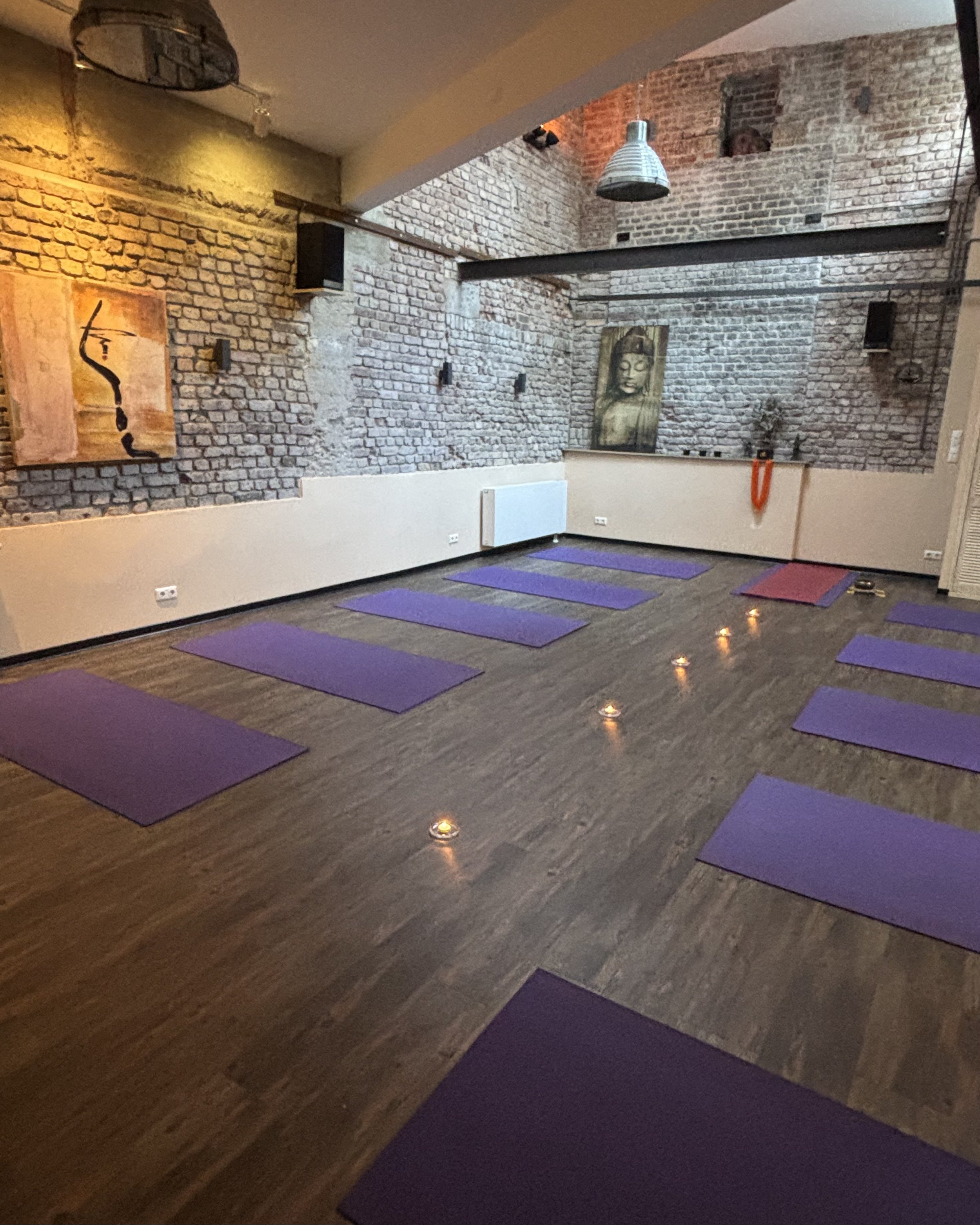 A welcoming and calming Yoga studio with purple mats on wooden floor, brick walls, artwork from Bali and candles on the floor, with a high ceiling and hanging lights.