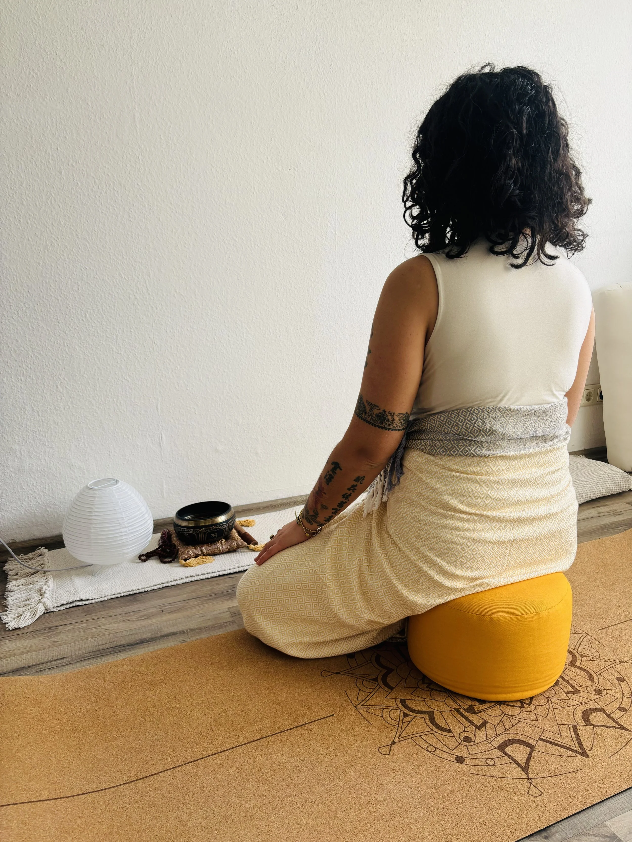 A woman sitting on a yellow meditation cushion on yoga mat, facing a wall with a small altar featuring a white lantern, a Tibetan sound bowl, practicing mindfulness in a minimalistic room.