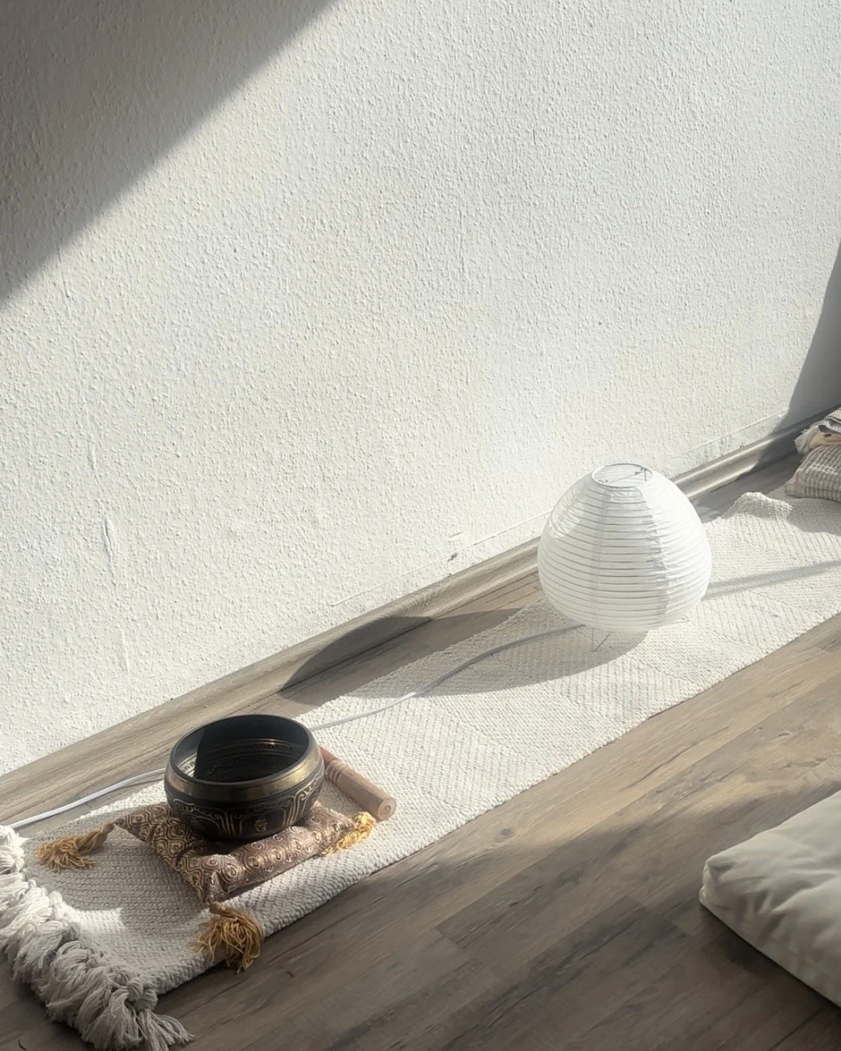 A white paper lantern on a wooden floor next to a Tibetan sound bowl on a decorative cloth, with a white textured wall and sunlight coming in the room.