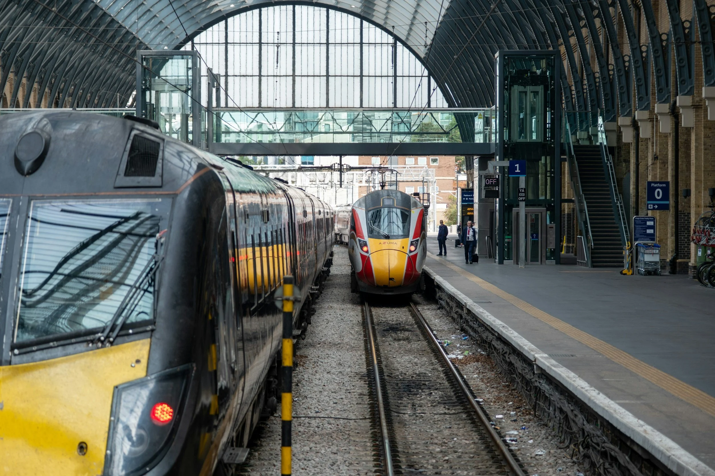 A modern train station with two trains on parallel tracks under a glass and metal arched roof. One train is black and yellow, the other is white and red. There are people walking and standing on the platform, and glass elevators and stairs are visible.
