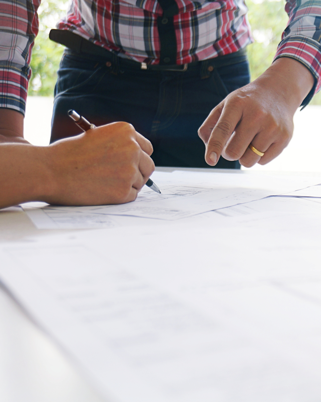 Person in plaid shirt and dark jeans pointing at blueprints while another person signs them.
