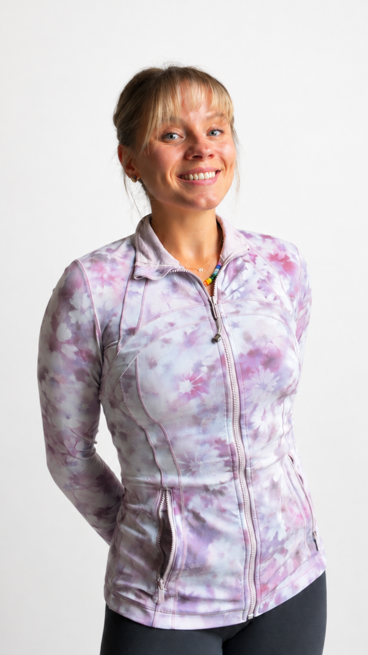 A woman who is a pilates instructor with blonde hair in a ponytail wearing a pink and purple floral zip-up athletic jacket, standing against a white background, smiling slightly with her hands behind her back.